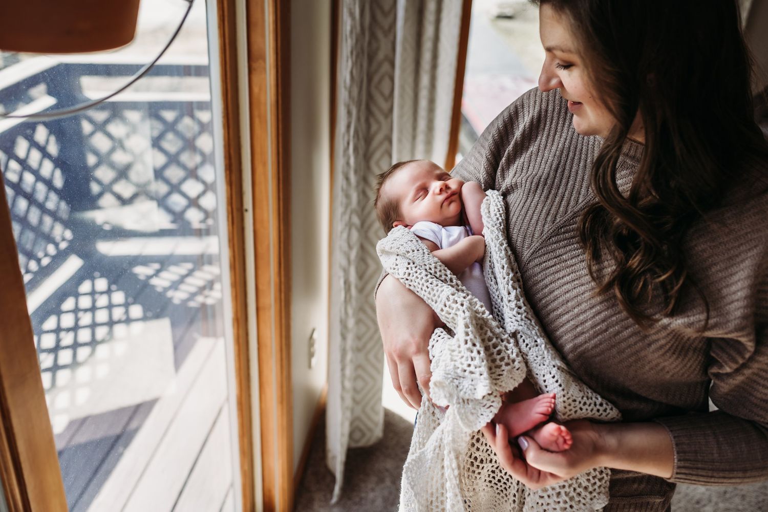 Bright window light illuminates baby wrapped in knit blanket by decorative curtains.