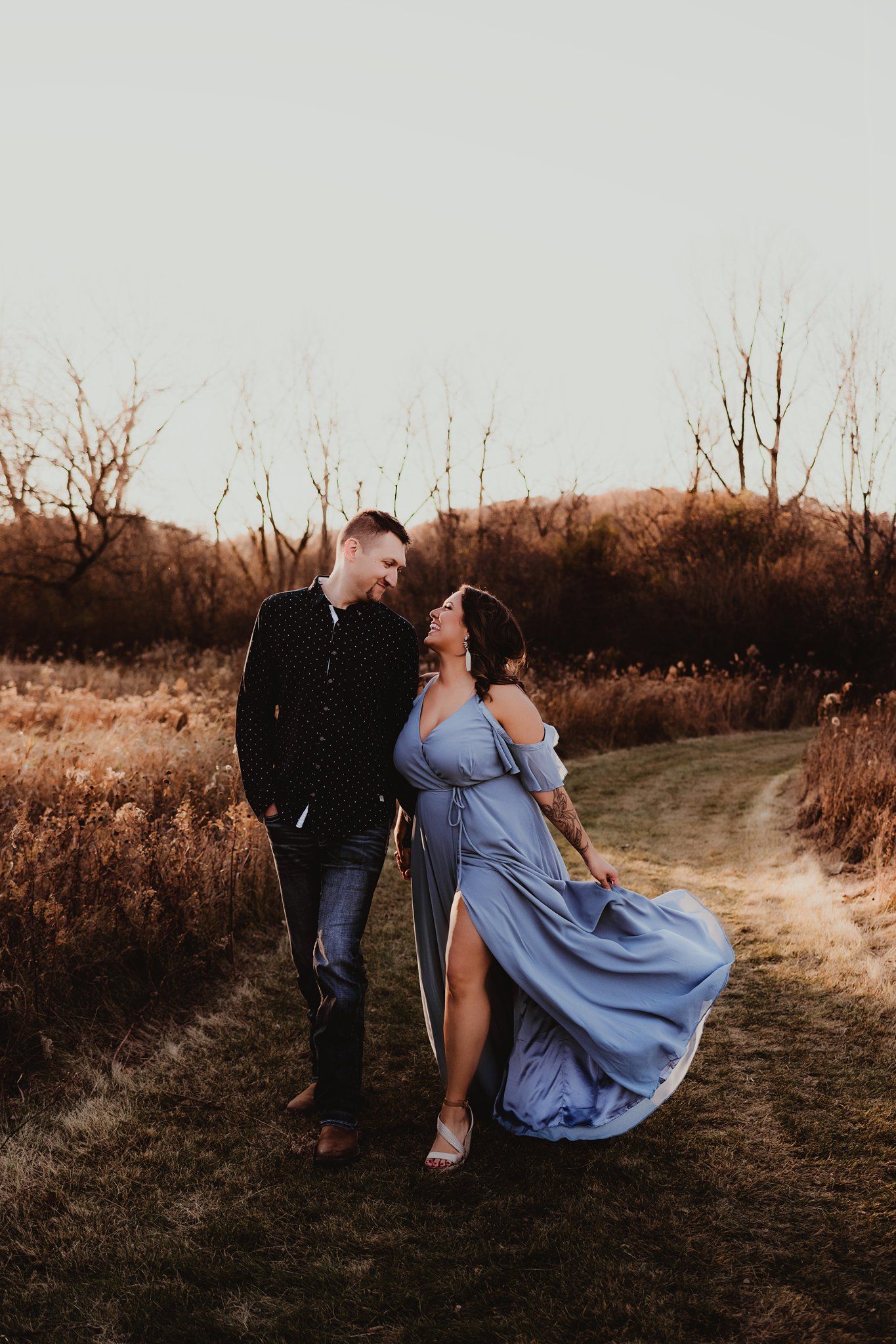 Elegant couple on dirt path with flowing blue dress catching wind at sunset.