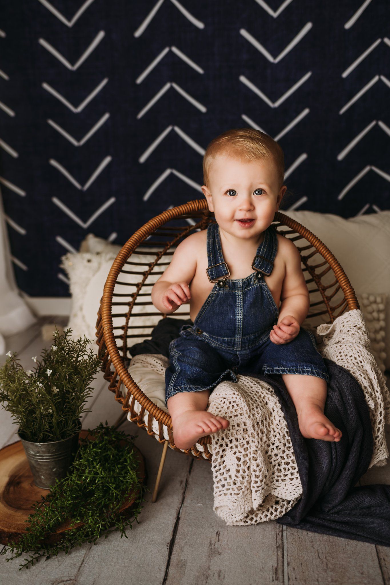 Smiling baby in denim overalls sits in rattan chair with knit blanket and greenery.