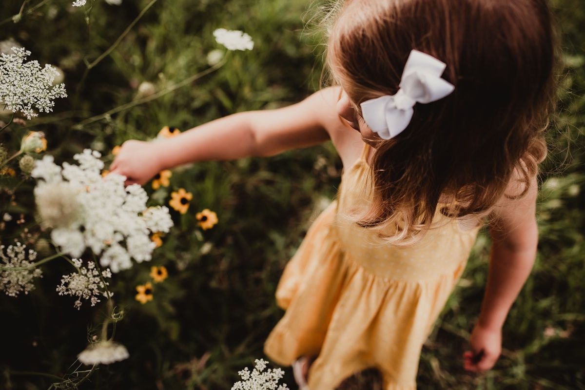 Child in a yellow dress reaching to touch white flowers in a garden.