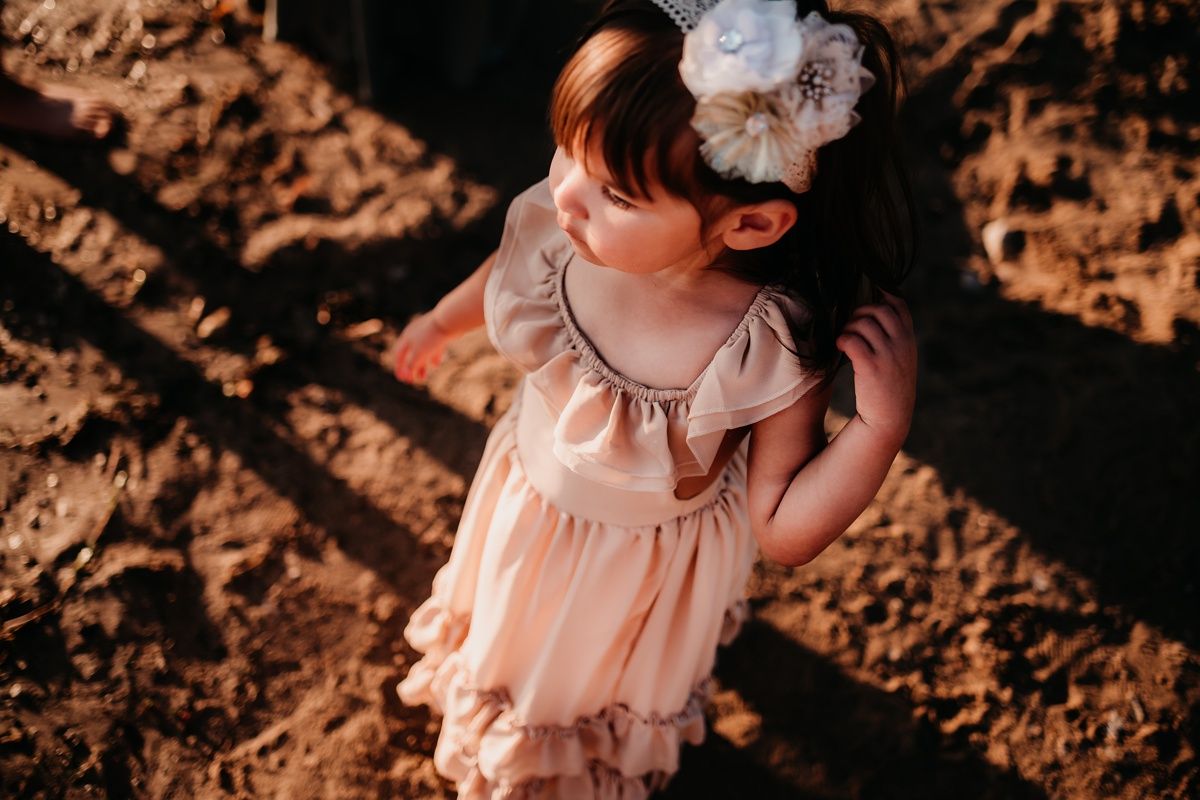 A small child wearing a peach-colored dress with ruffles stands in warm sunlight outdoors.