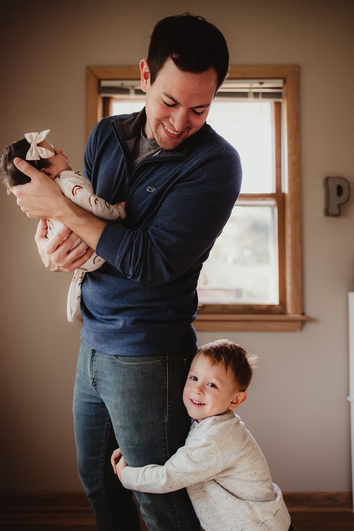 Parent holding infant while young child hugs their legs in sunlit room.
