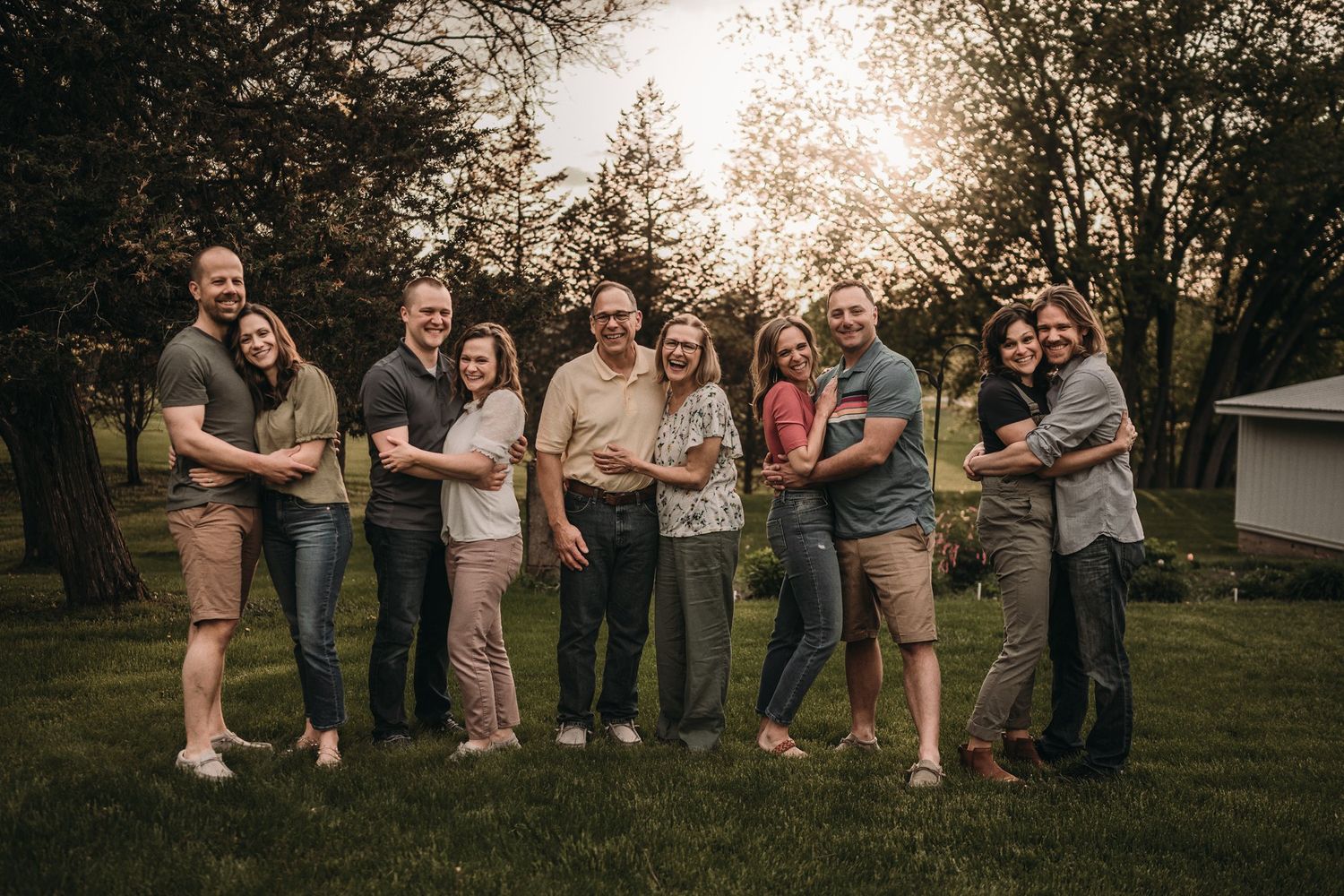 Multiple couples stand together embracing in a park during sunset.
