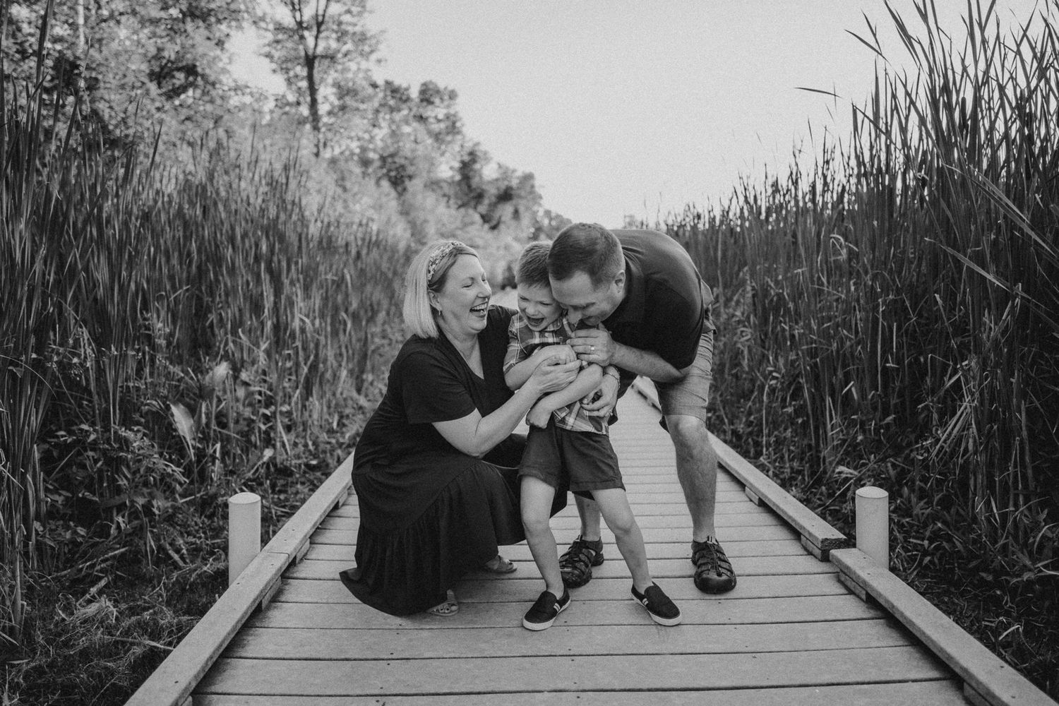 A tender family moment captured on a wooden boardwalk surrounded by tall reeds in black and white.