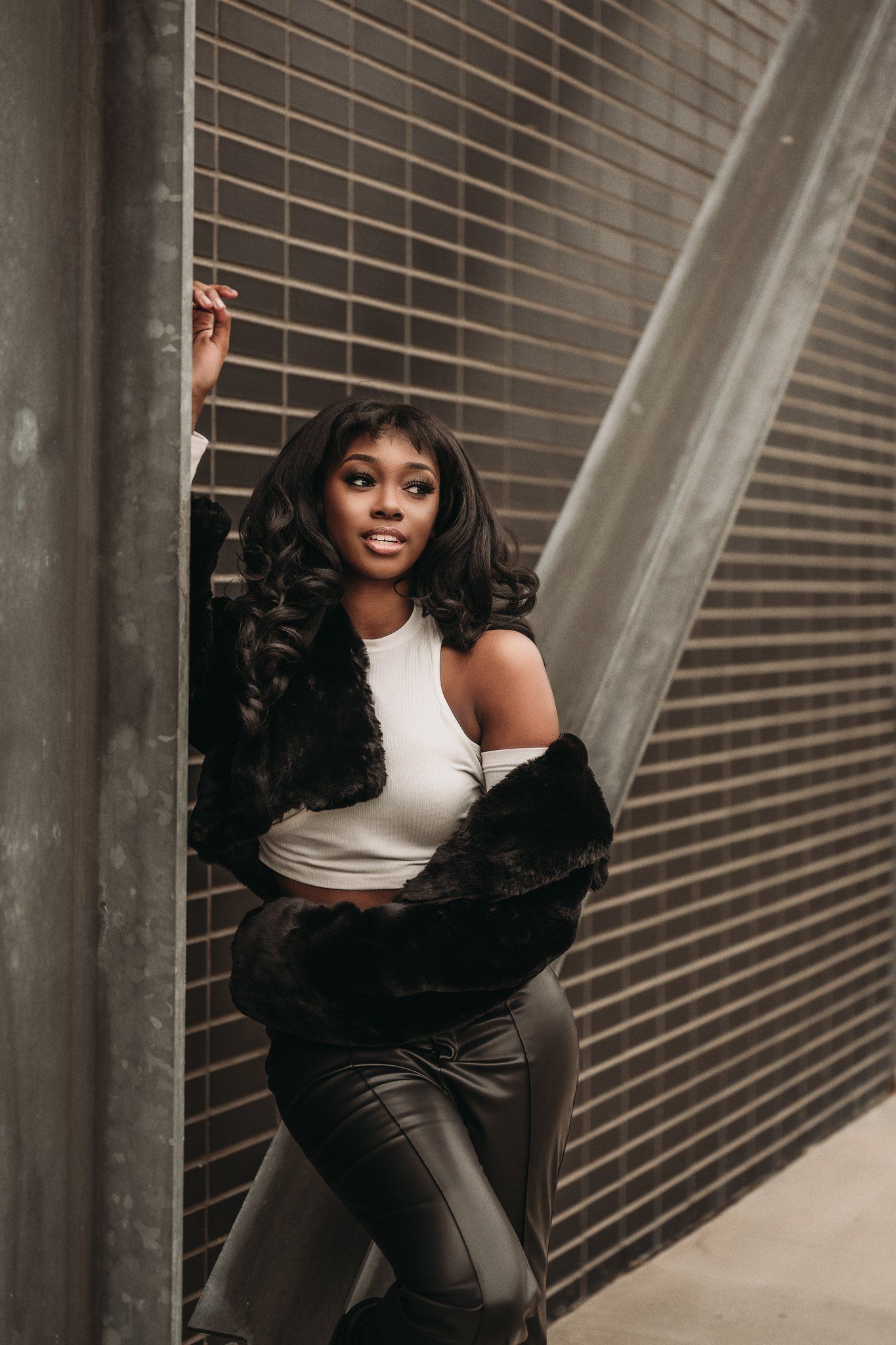 Model in white crop top and faux fur coat poses dramatically by industrial wall.
