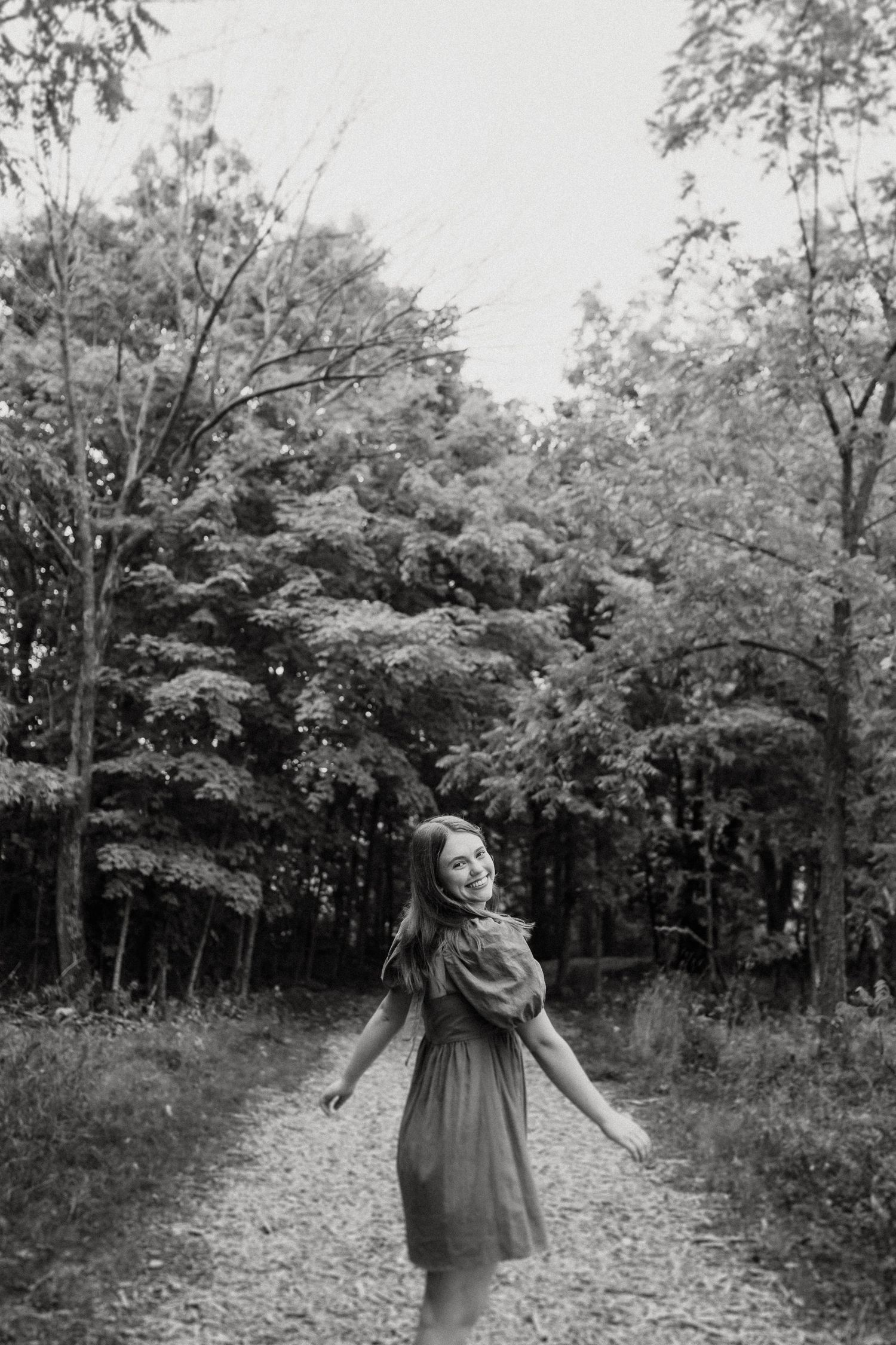 Black and white image of someone twirling in a dress on a wooded path.