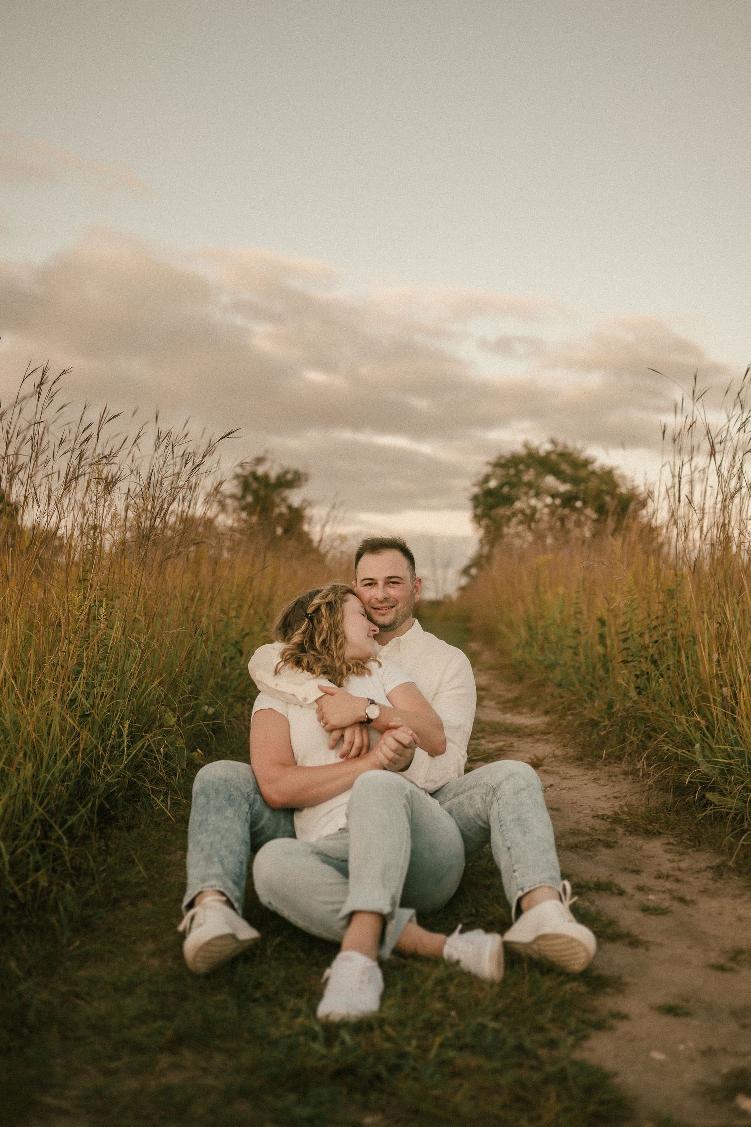 Couple sits together on dirt path surrounded by tall grasses at dusk.