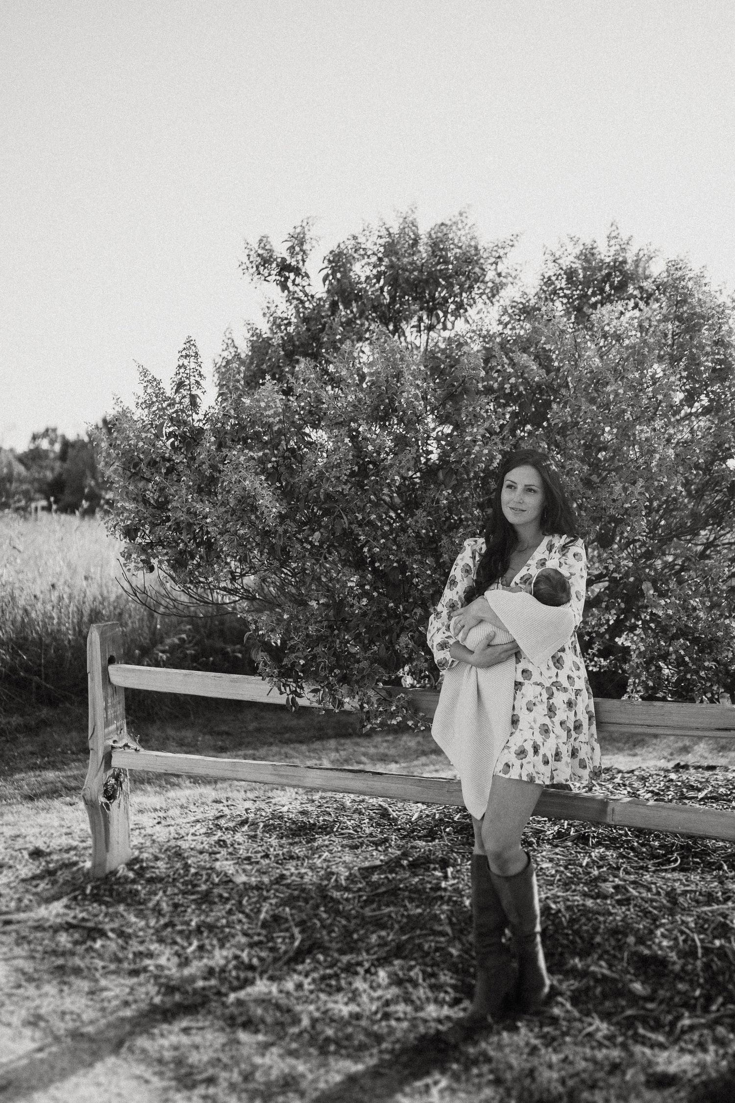 Woman in floral dress stands by rustic wooden fence near flowering bush.