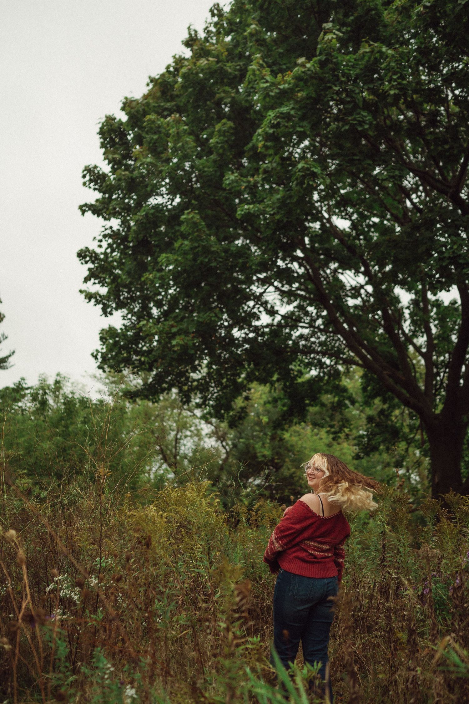 Person in a red sweater stands beneath a towering tree in a wild meadow setting.