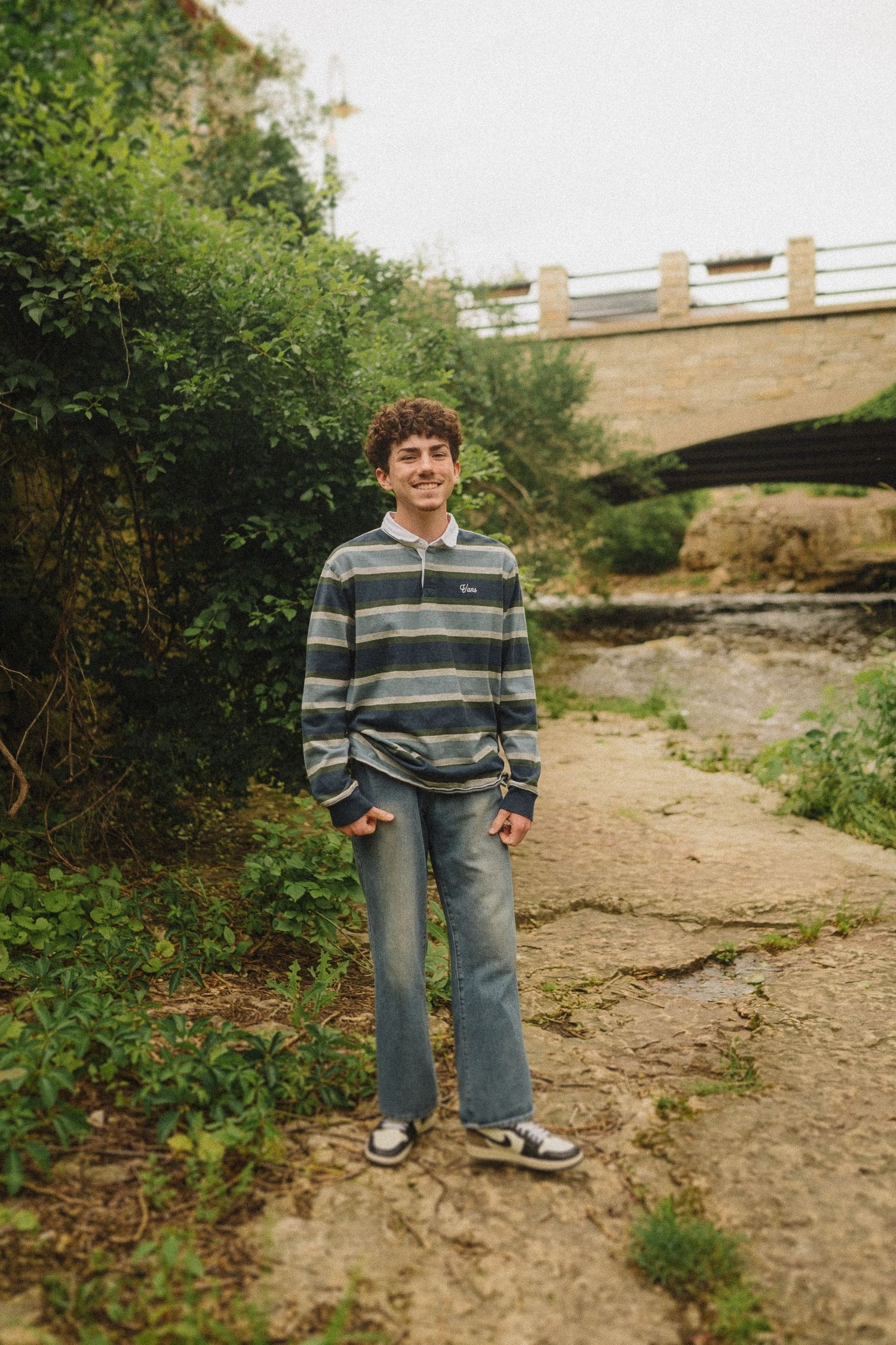 A person in a striped pullover stands on a dirt path beneath a stone bridge in a wooded area.