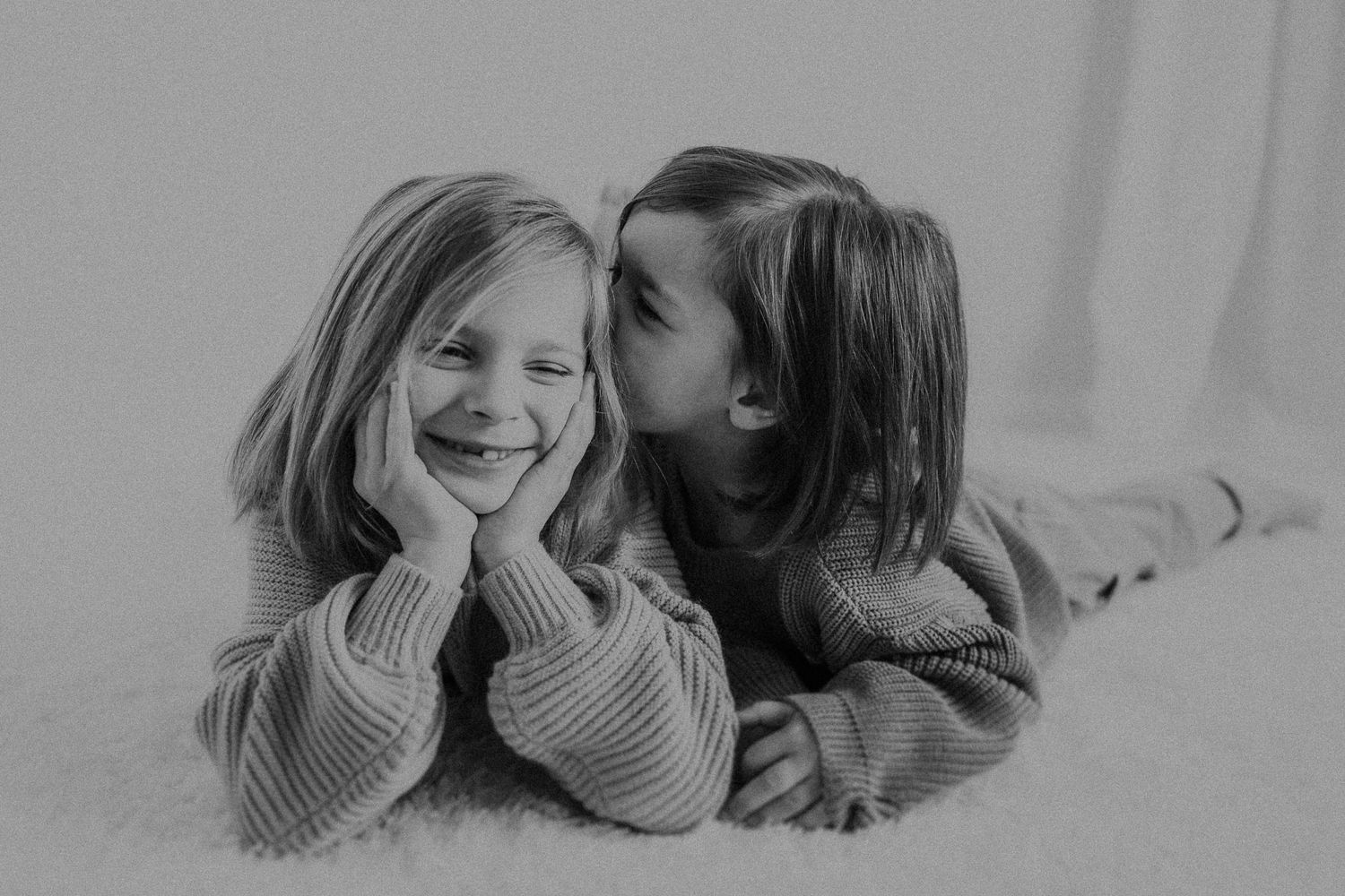 Two children share a sweet moment while lying on a light carpet in black and white.