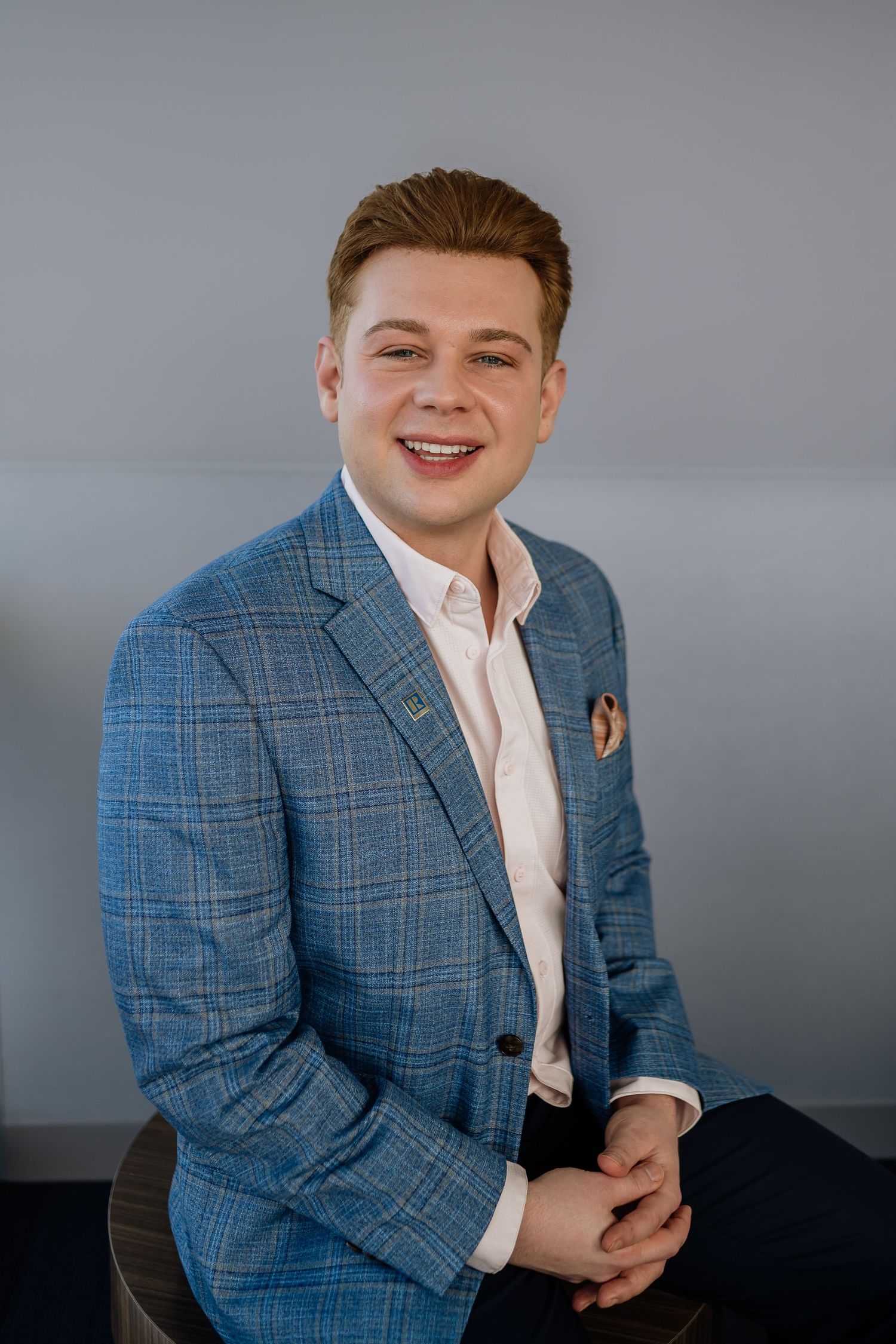 Professional headshot of person wearing blue plaid blazer against grey background.