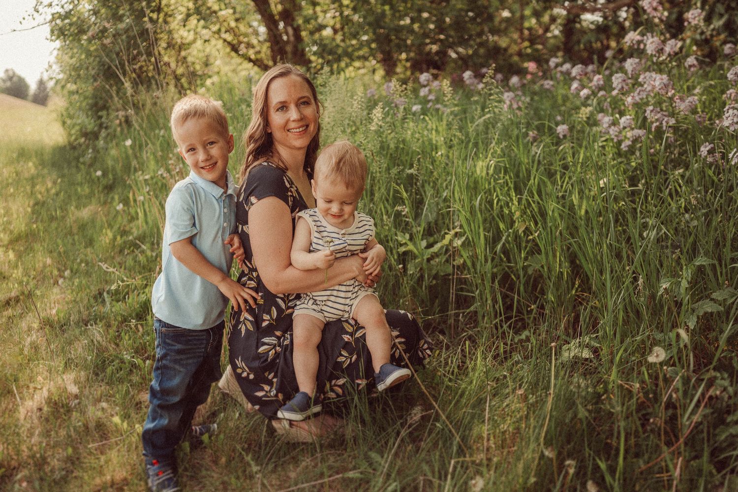 A warm family portrait outdoors with tall grass and wildflowers in golden sunlight.