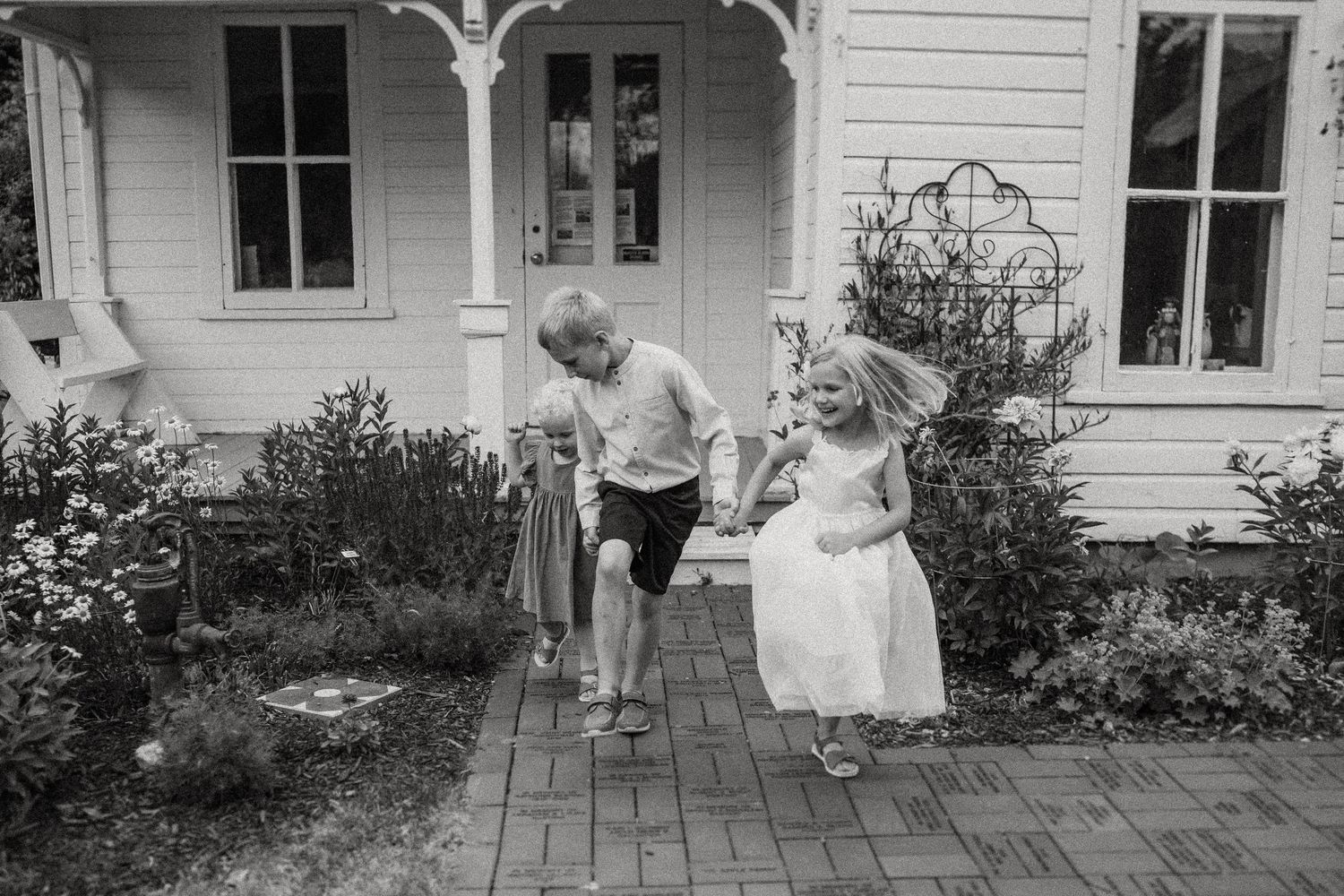 Black and white photo of children playing and running on brick pathway by farmhouse.