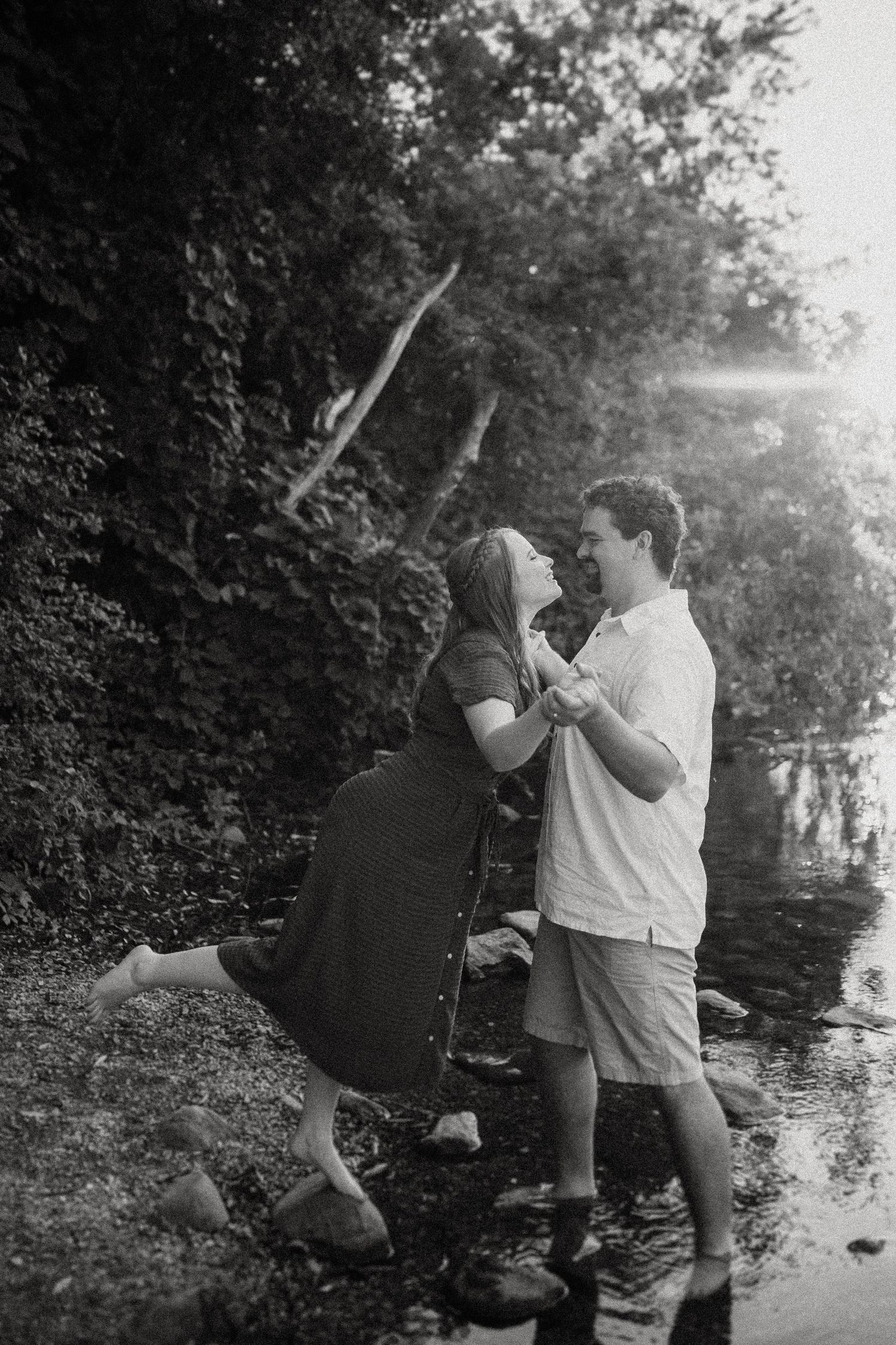 Black and white romantic photo of a couple by the water's edge with trees in the background.