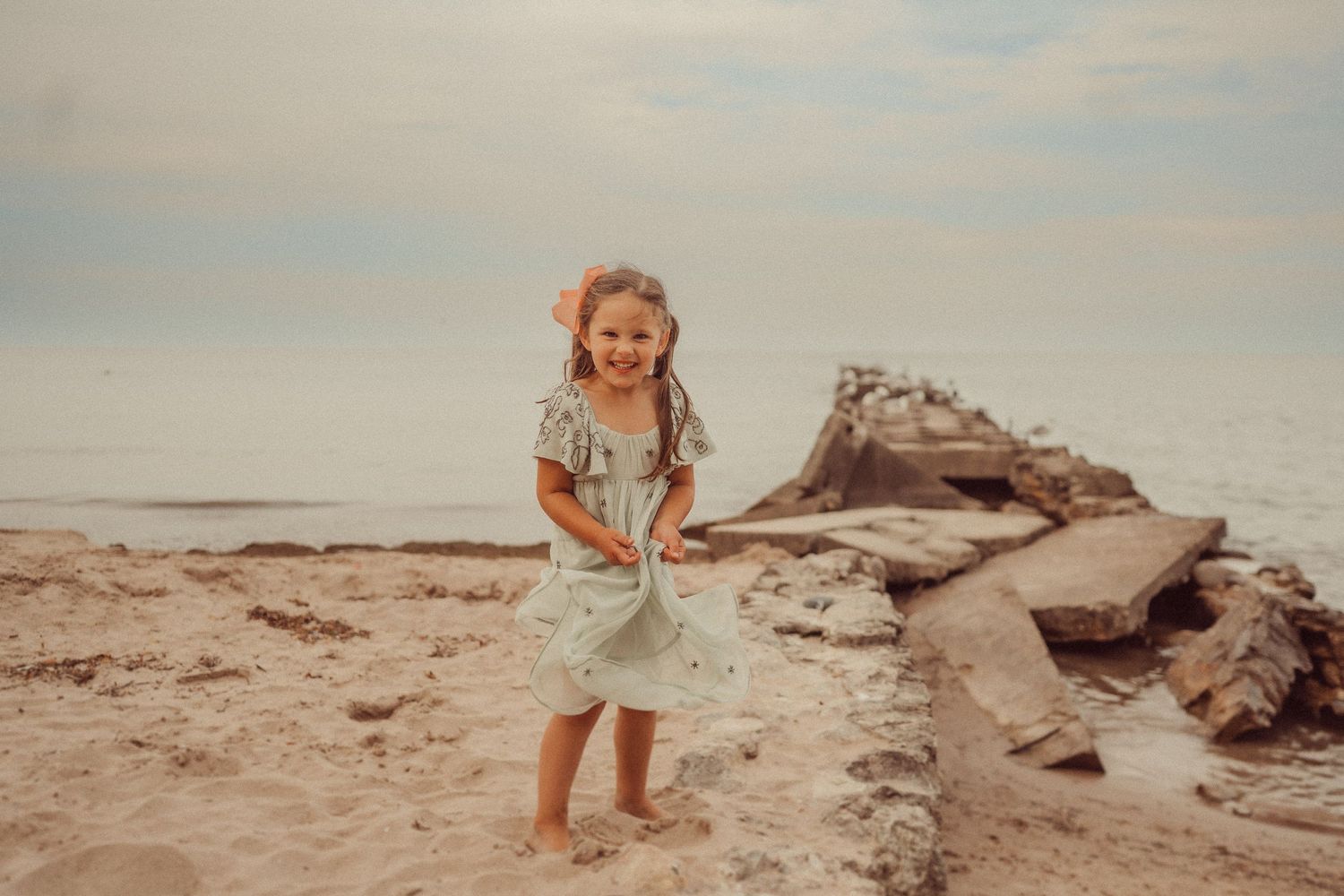 Young child in light dress plays happily on rocky beach during sunset.