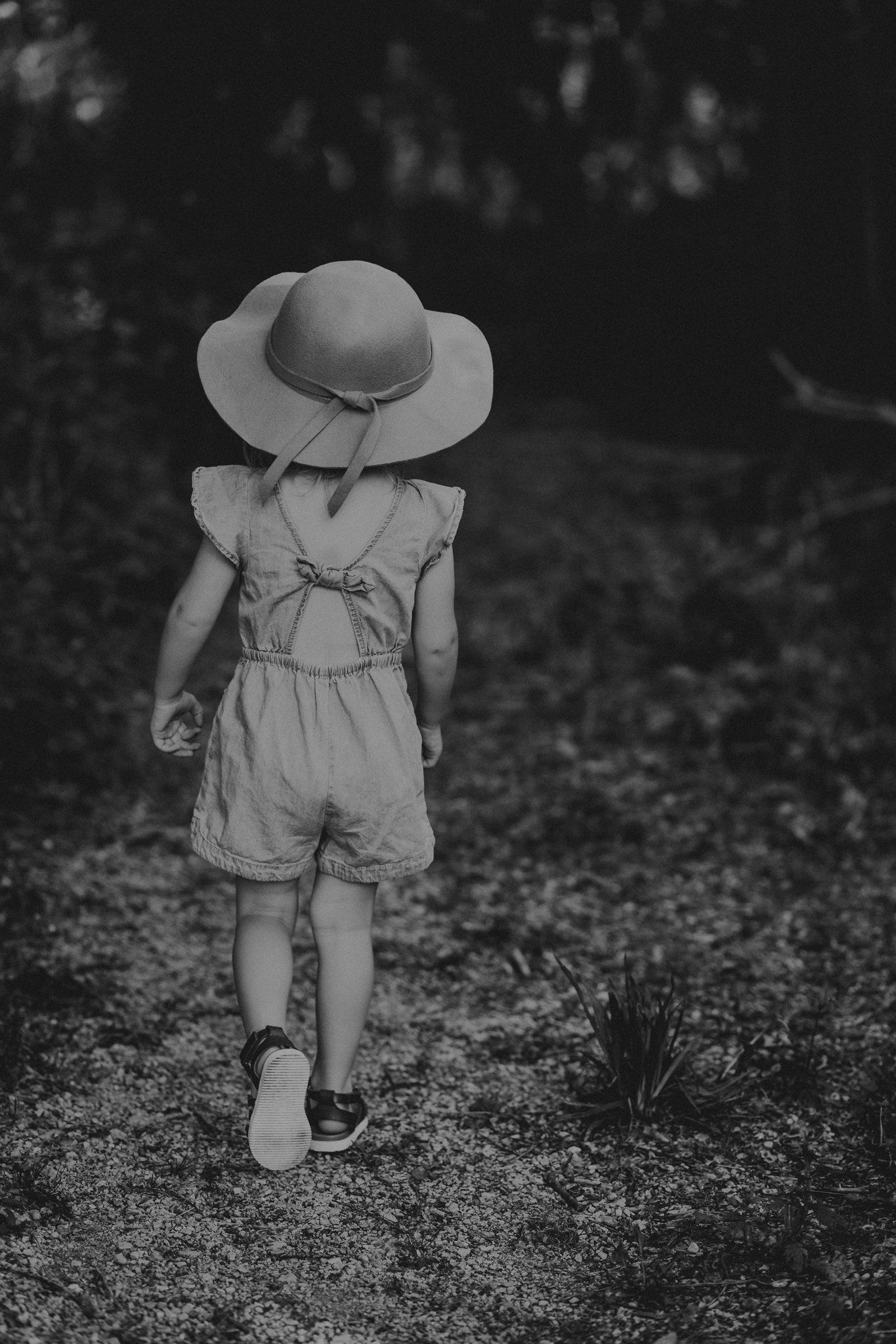 Back view of a child in a romper and sun hat walking along a wooded path.