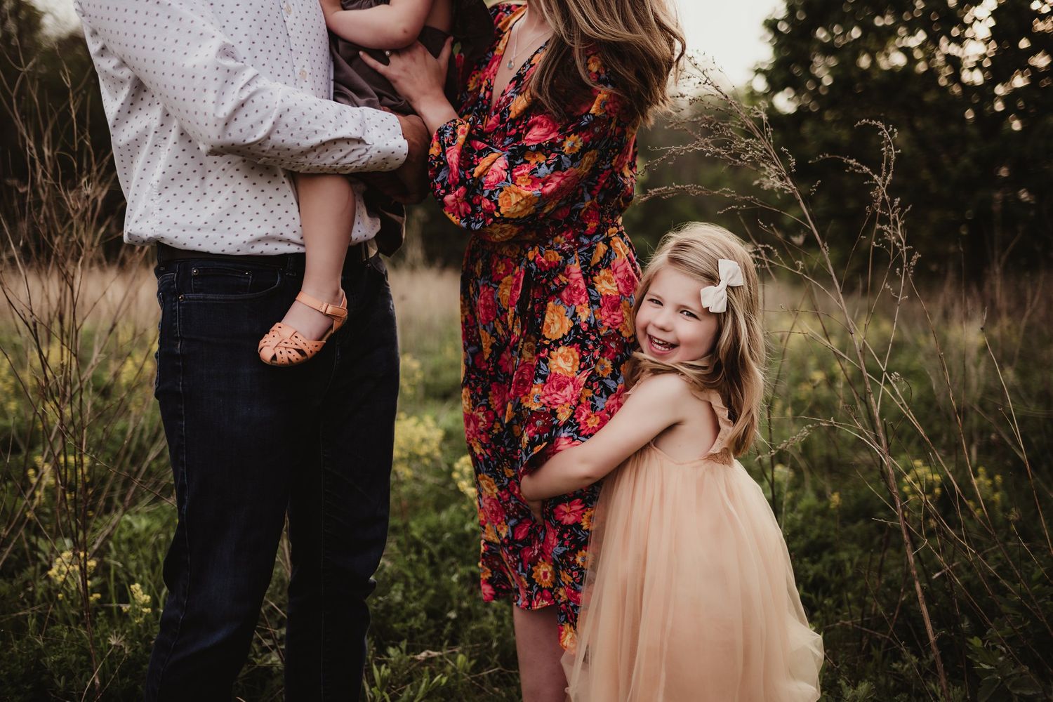 A family embraces in a golden field at sunset wearing coordinated outfits of floral dress and neutral tones.