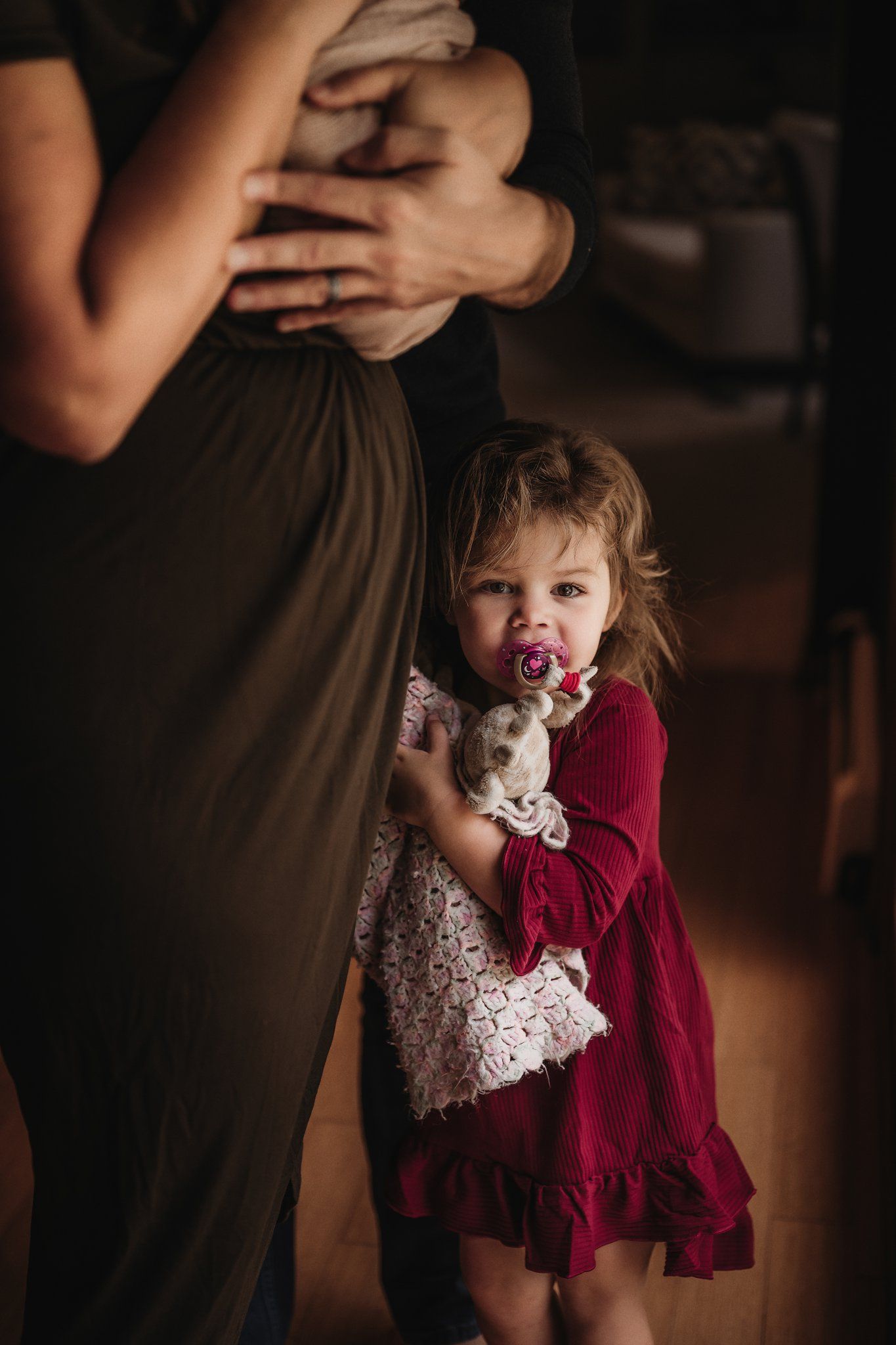 Child in red dress holding stuffed toy while standing next to parent holding baby.