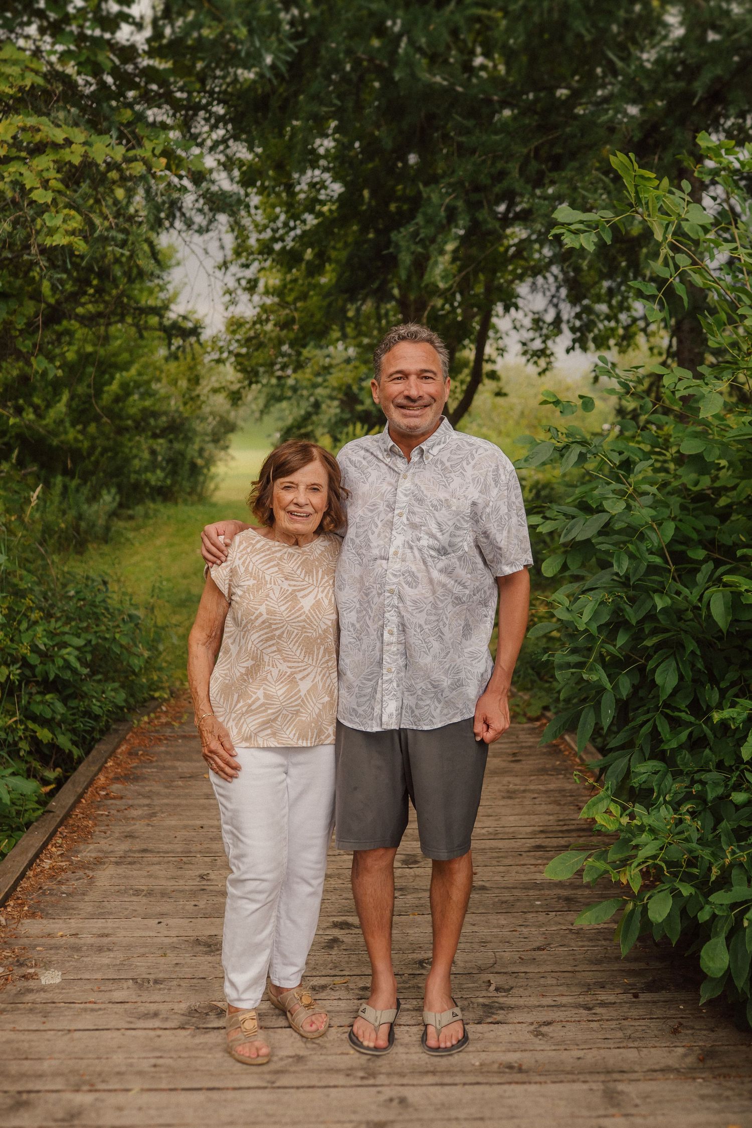A couple poses together on a wooden footbridge surrounded by lush green foliage in summer.
