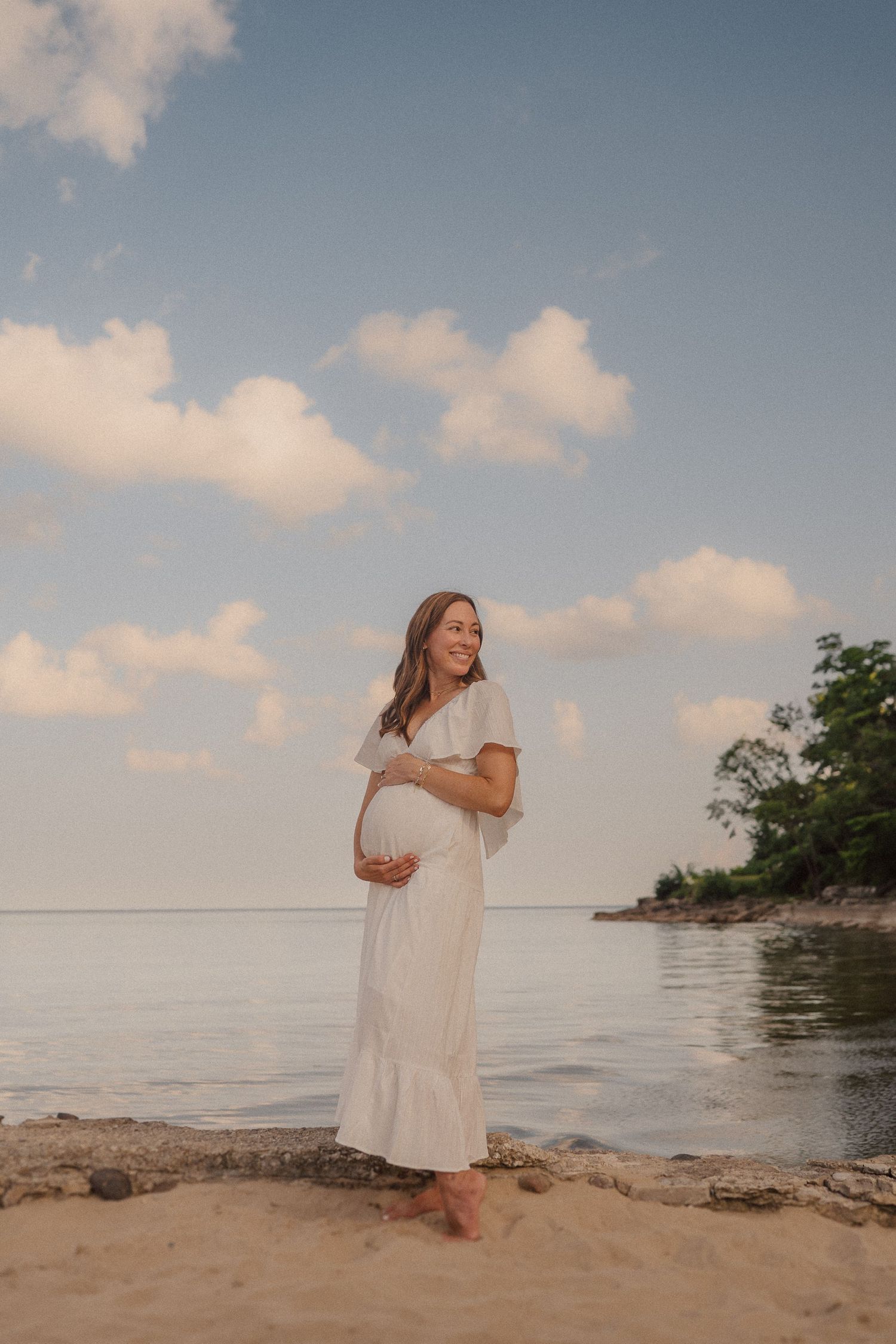 A woman in a white dress stands on a beach during sunset with clouds and water in the background.