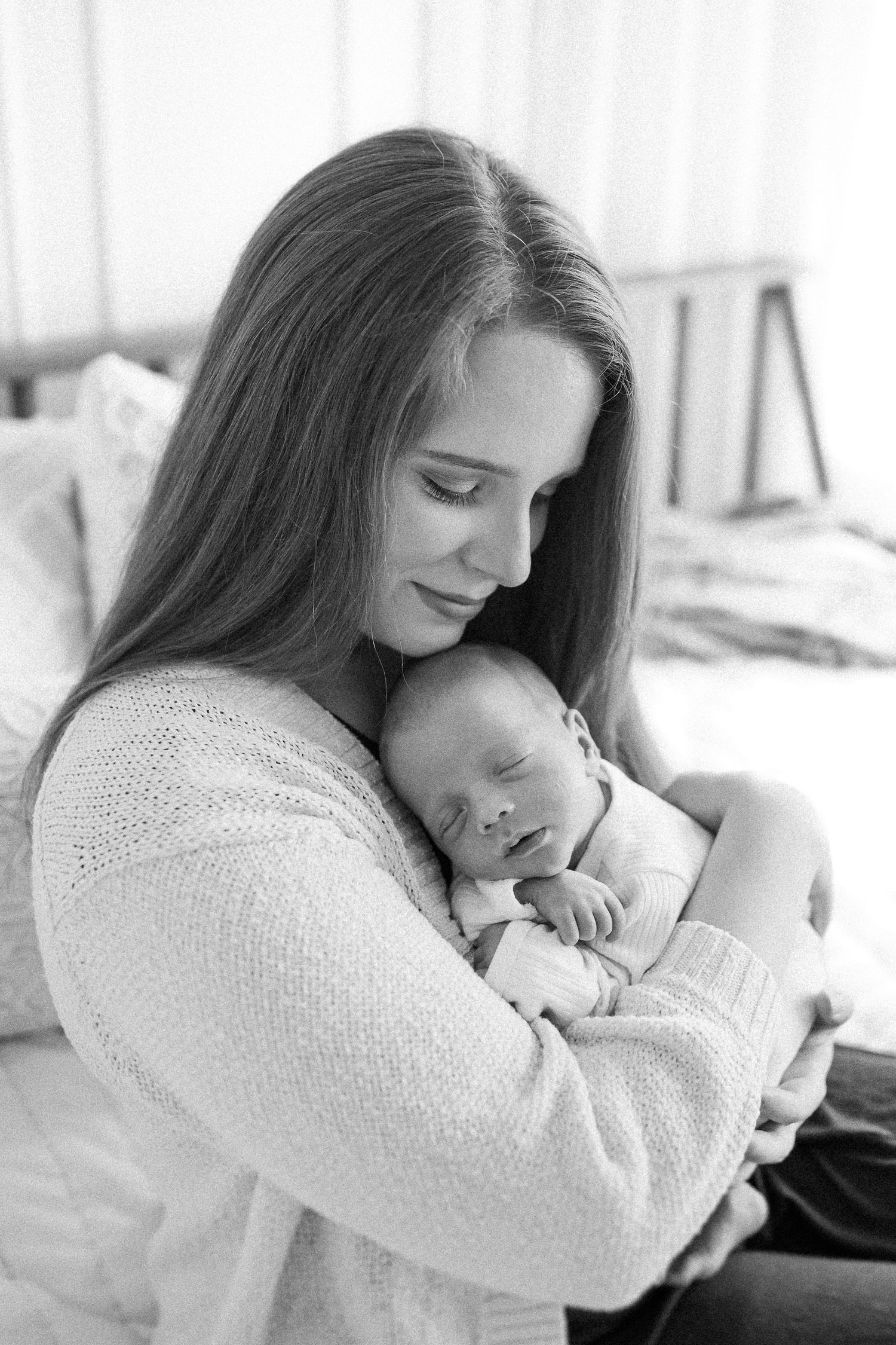 A tender black and white photo of a mother in a sweater cradling her sleeping newborn baby against her chest.