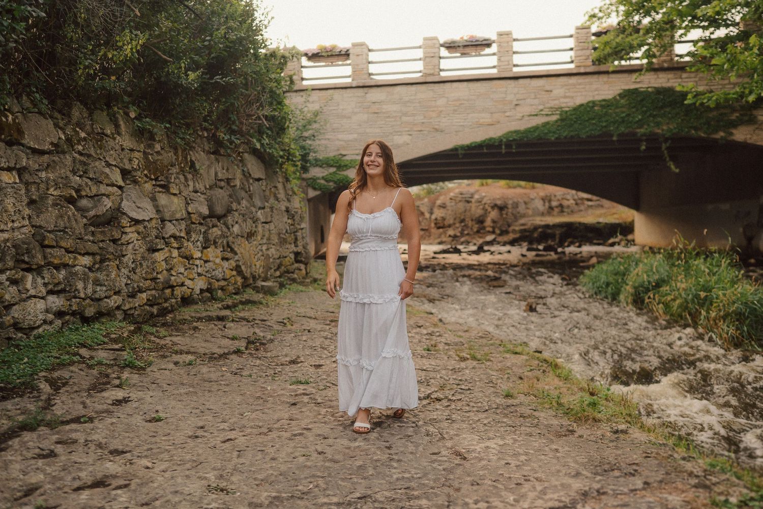 A person in a flowing white dress stands on a dry creek bed beneath a stone bridge.