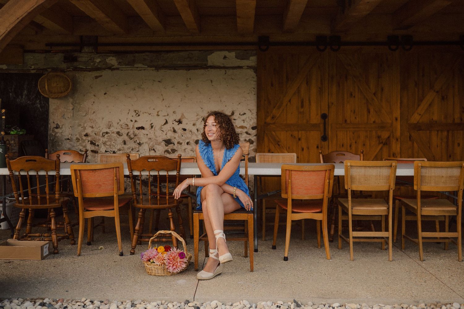 A rustic cafe scene with wooden chairs and a wicker basket sitting on a patio against a weathered stone wall.