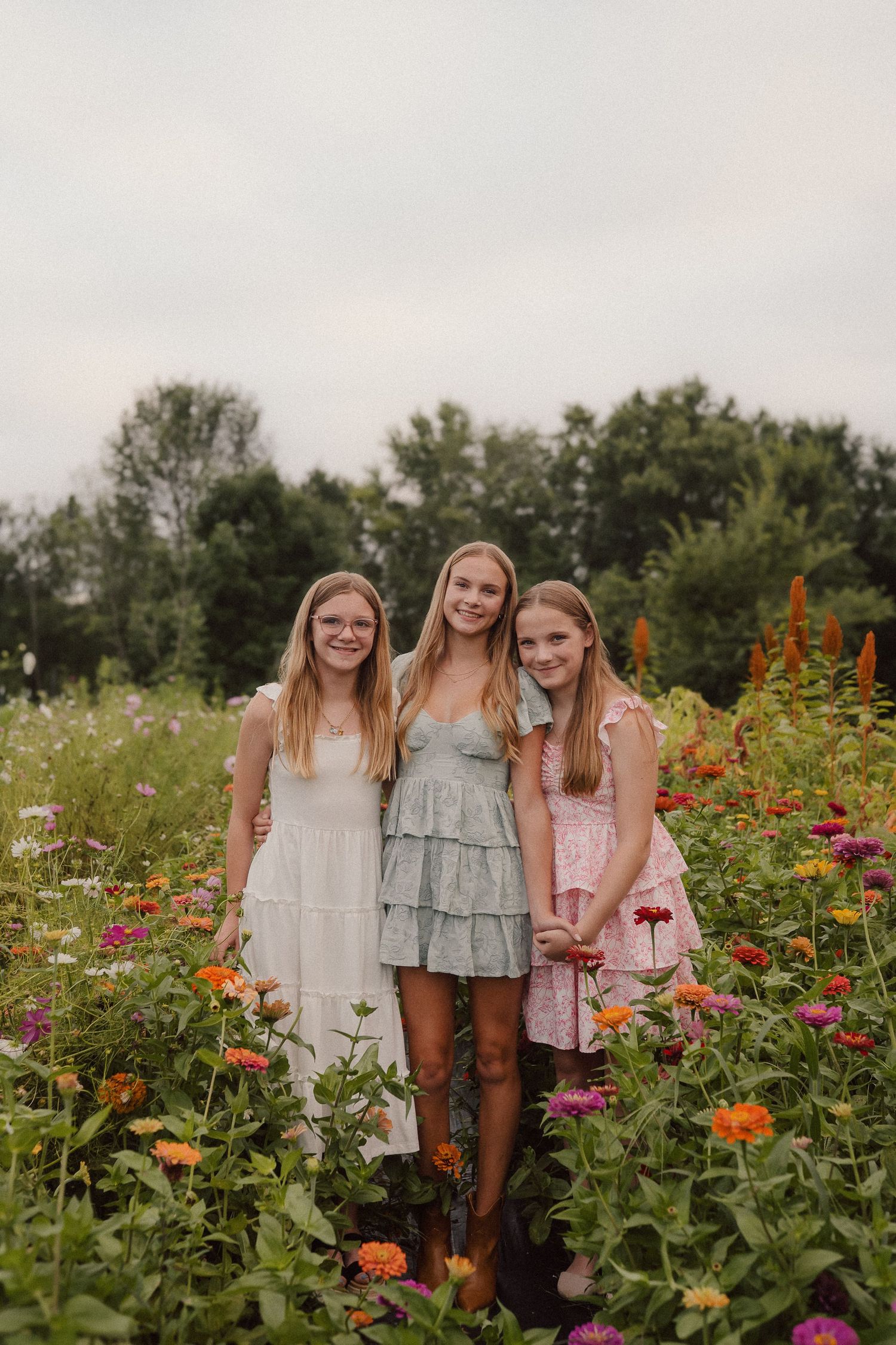 Three friends stand together in a blooming summer flower garden filled with colorful zinnias and wildflowers.