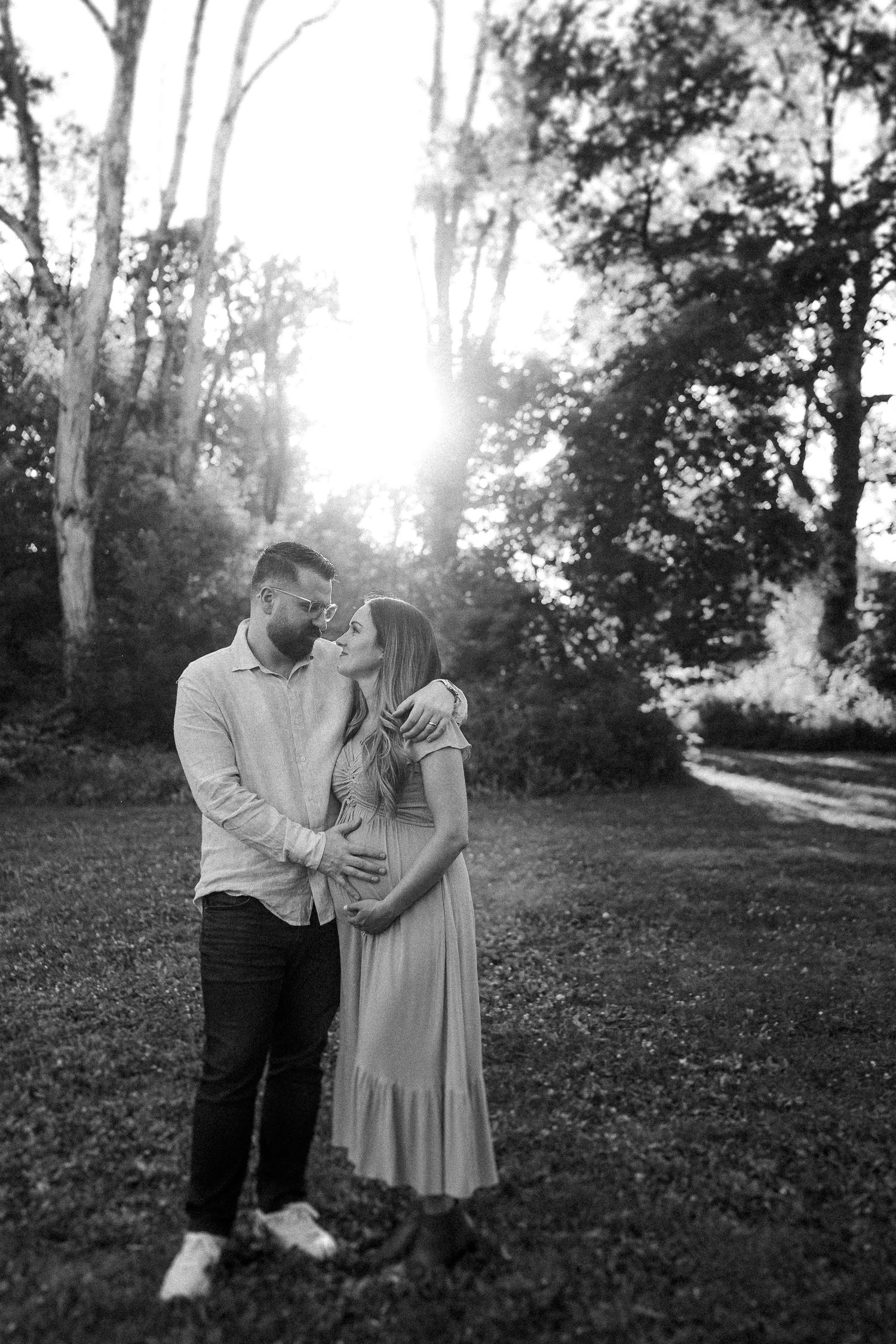 A romantic outdoor photo session shows a couple embracing in a sunlit wooded area wearing light colored clothing.