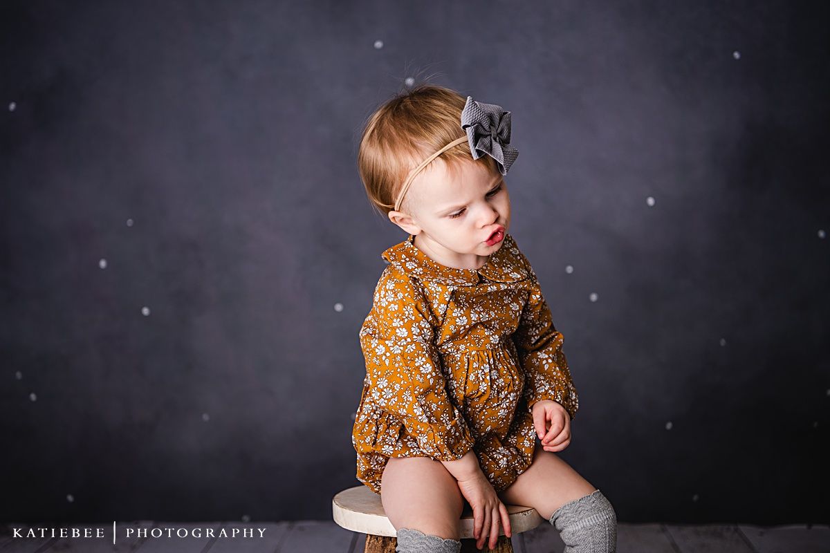 Child wearing mustard floral romper and bow headband posing on wooden stool.