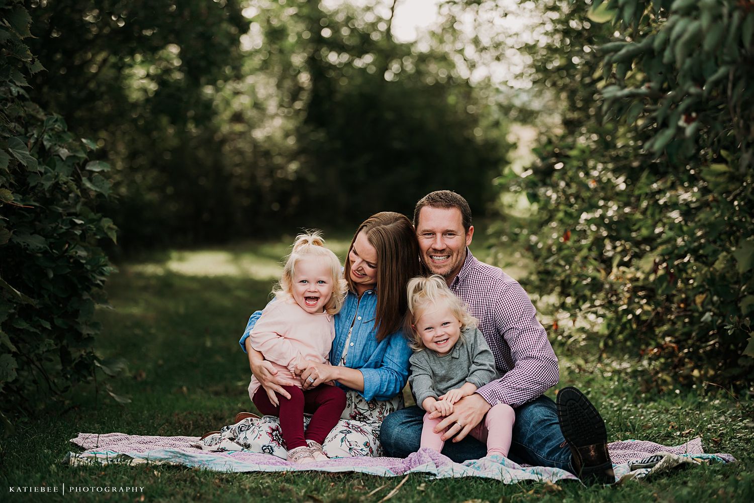 Family shares a candid moment of laughter while sitting on a blanket in a garden setting.