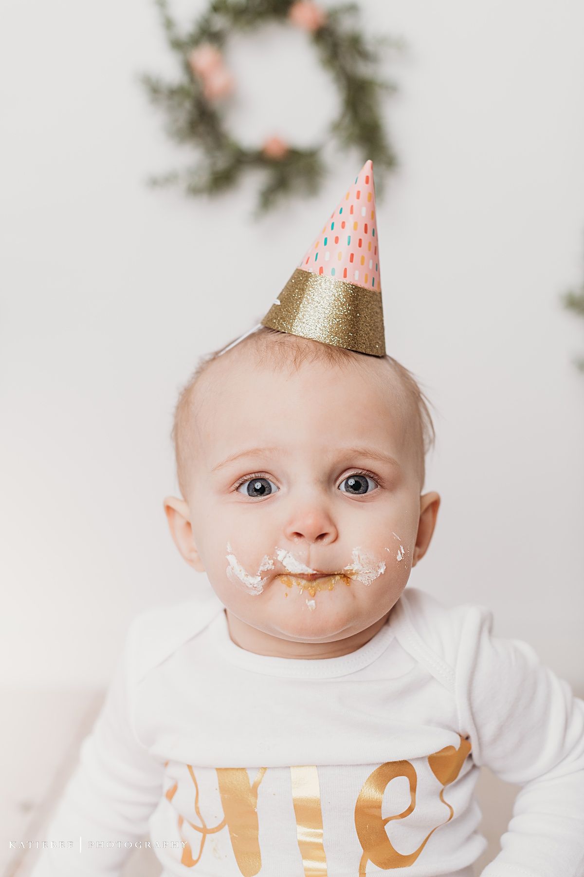 Baby wearing gold and pink party hat with cake frosting on face during first birthday celebration.