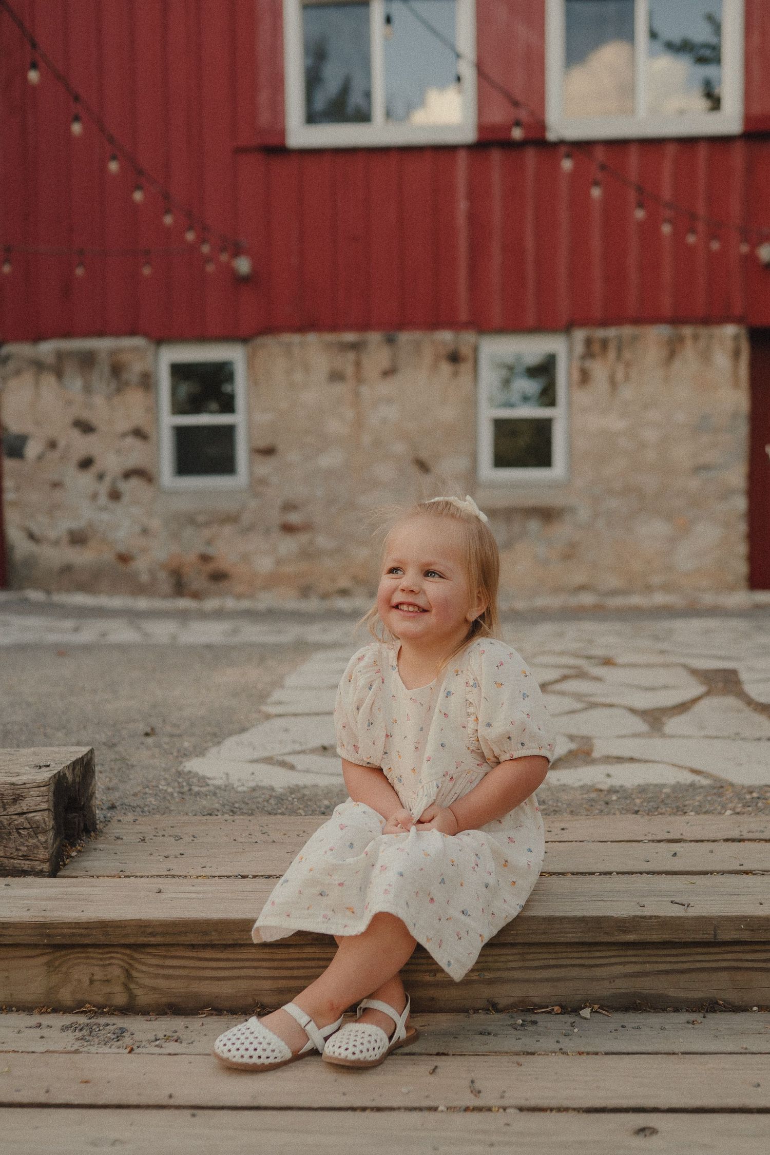 Happy toddler girl in white floral dress sitting on wooden steps smiling in front of a red barn.