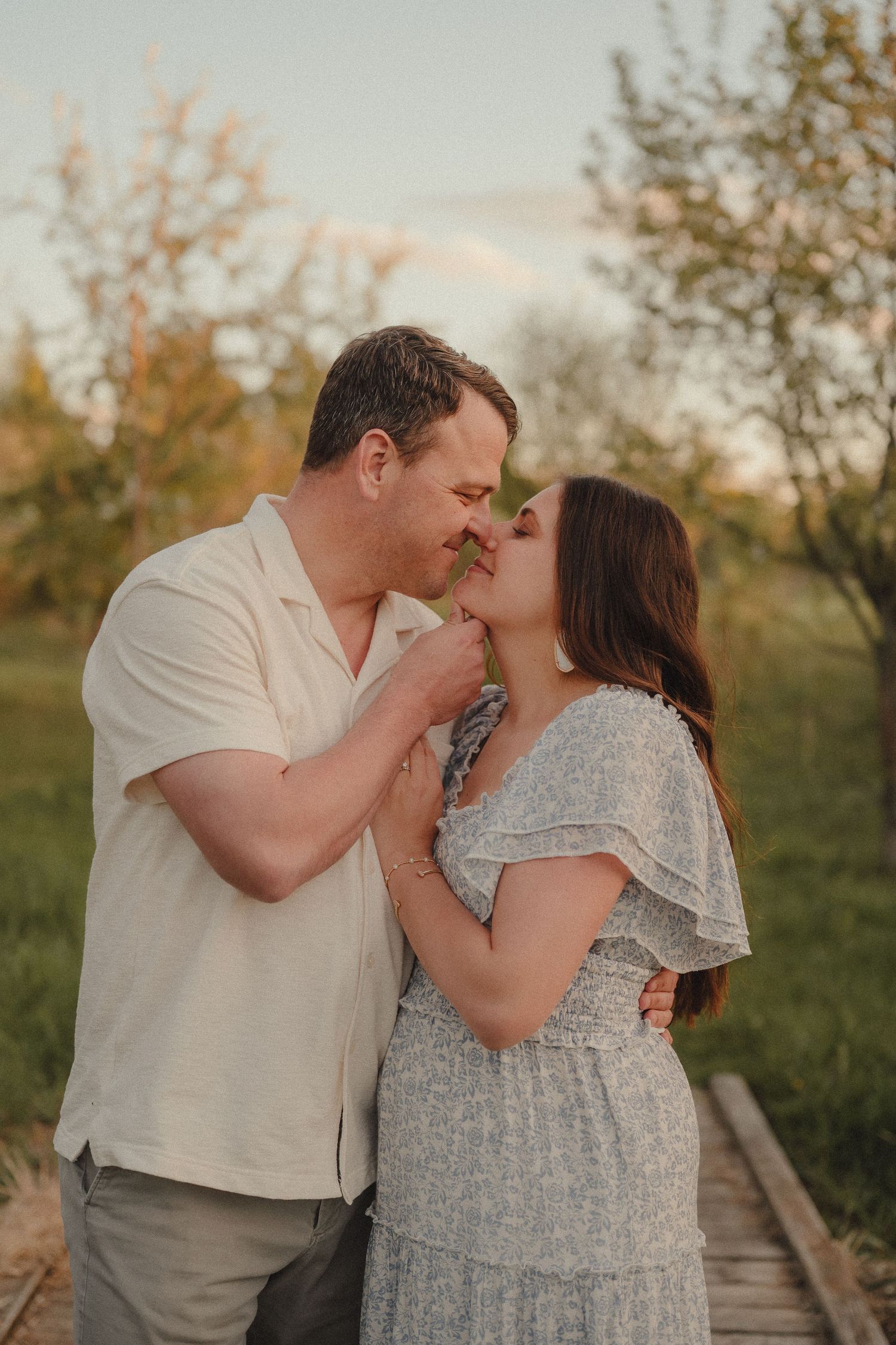 Romantic couple sharing a kiss on a wooden boardwalk path in a lush green orchard at sunset.