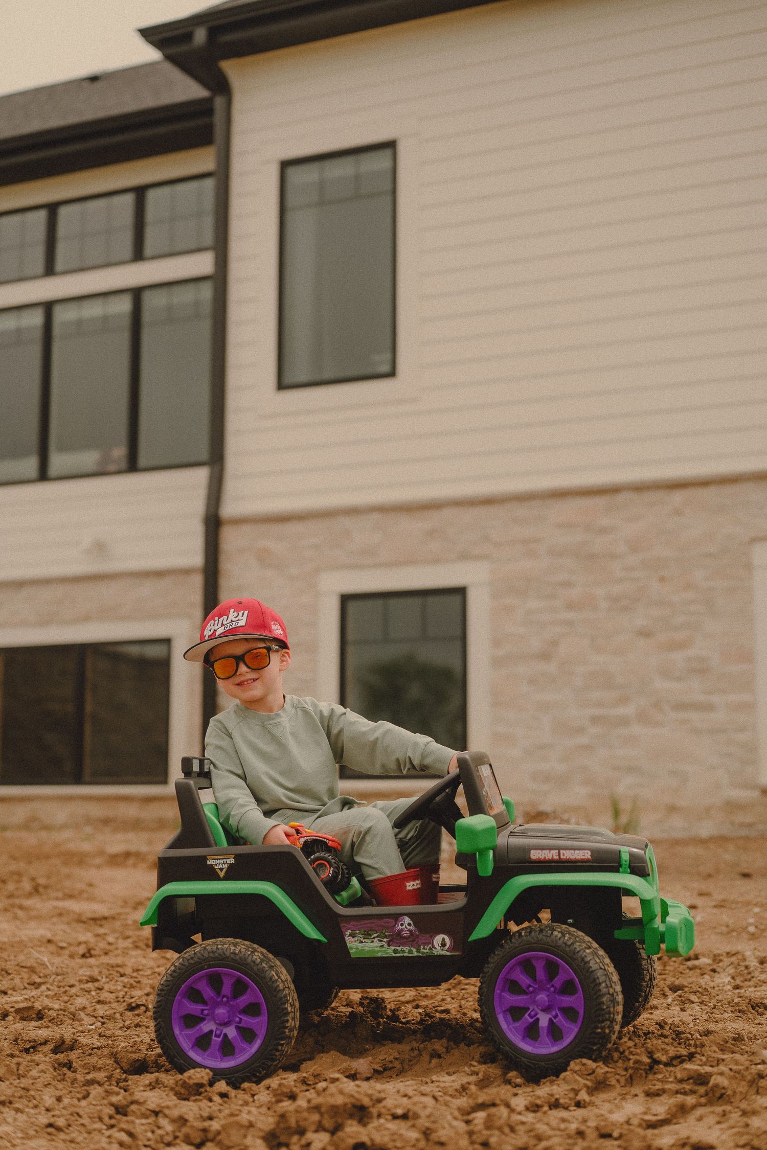 Young boy wearing sunglasses drives green ride-on toy jeep in dirt near modern house.