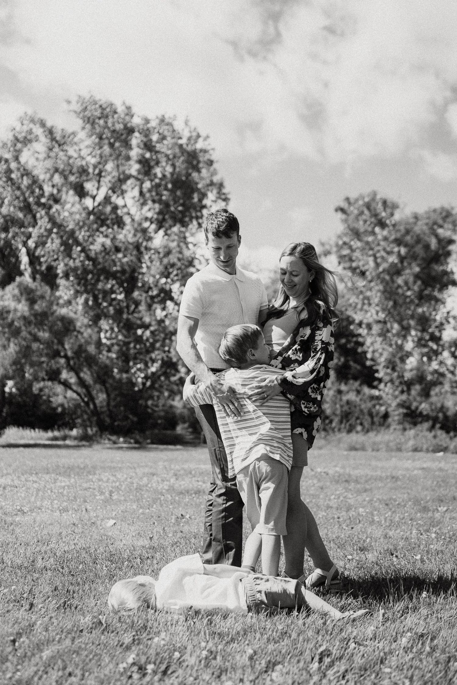 Black and white family photo of parents with young children playing together on a sunny day in a green park.