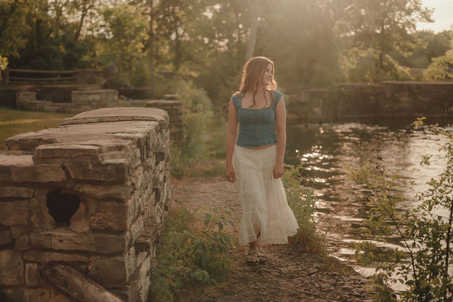 Woman in white skirt and teal top walking near a stone bridge by a sunlit river in a lush park.