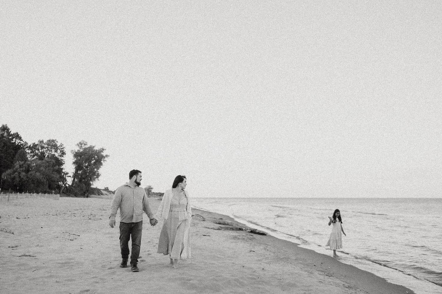 Black and white photo of a couple walking hand in hand on a beach with a child running ahead near the water.