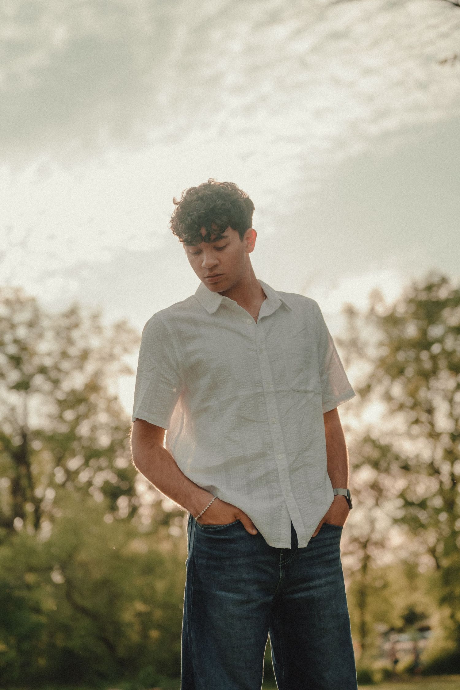 Young man with curly hair wearing white shirt and jeans standing outdoors under cloudy sky.