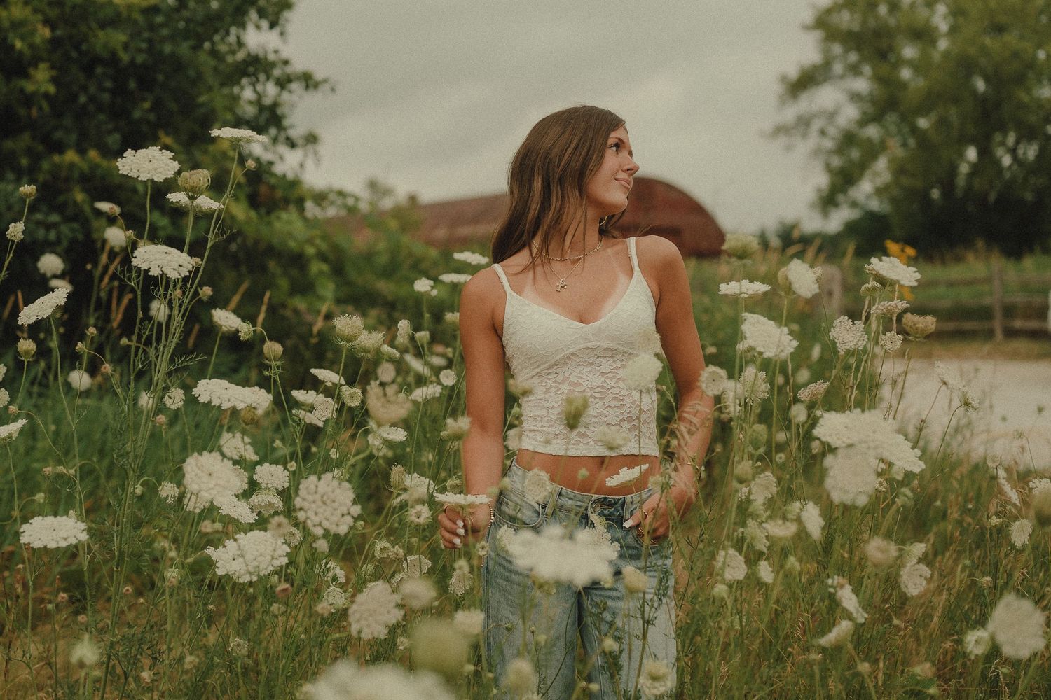 Young woman in white lace crop top and jeans standing in a field of white wildflowers on a cloudy day.