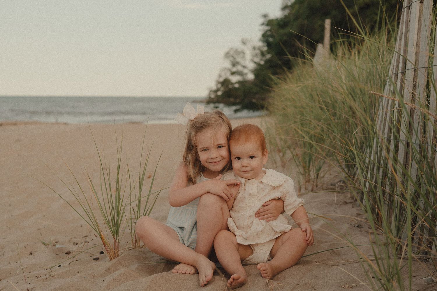 Two young siblings smile together on a sandy beach, with ocean waves and green dune grass in the background.