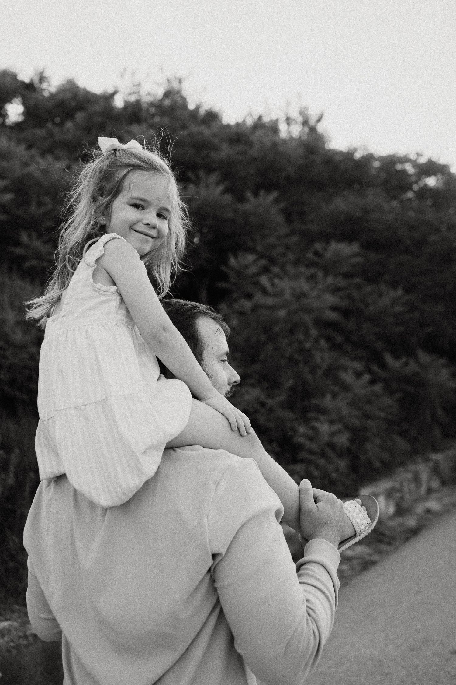 Black and white photo of a smiling little girl sitting on a man's shoulders outdoors near lush green trees.
