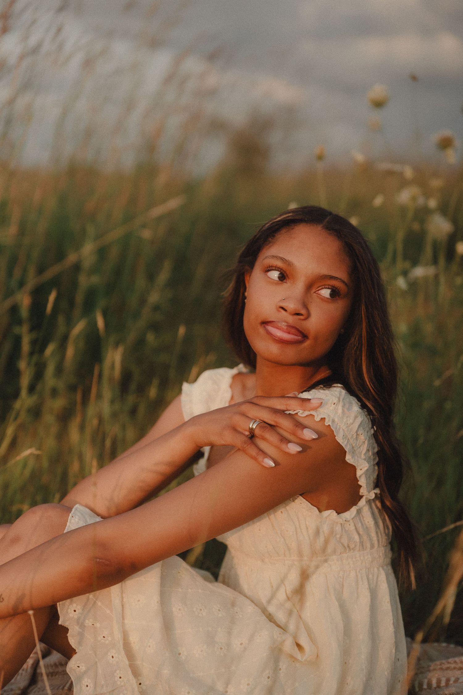 Young woman in a white eyelet dress sits in a golden field at dusk, gazing thoughtfully into the distance.