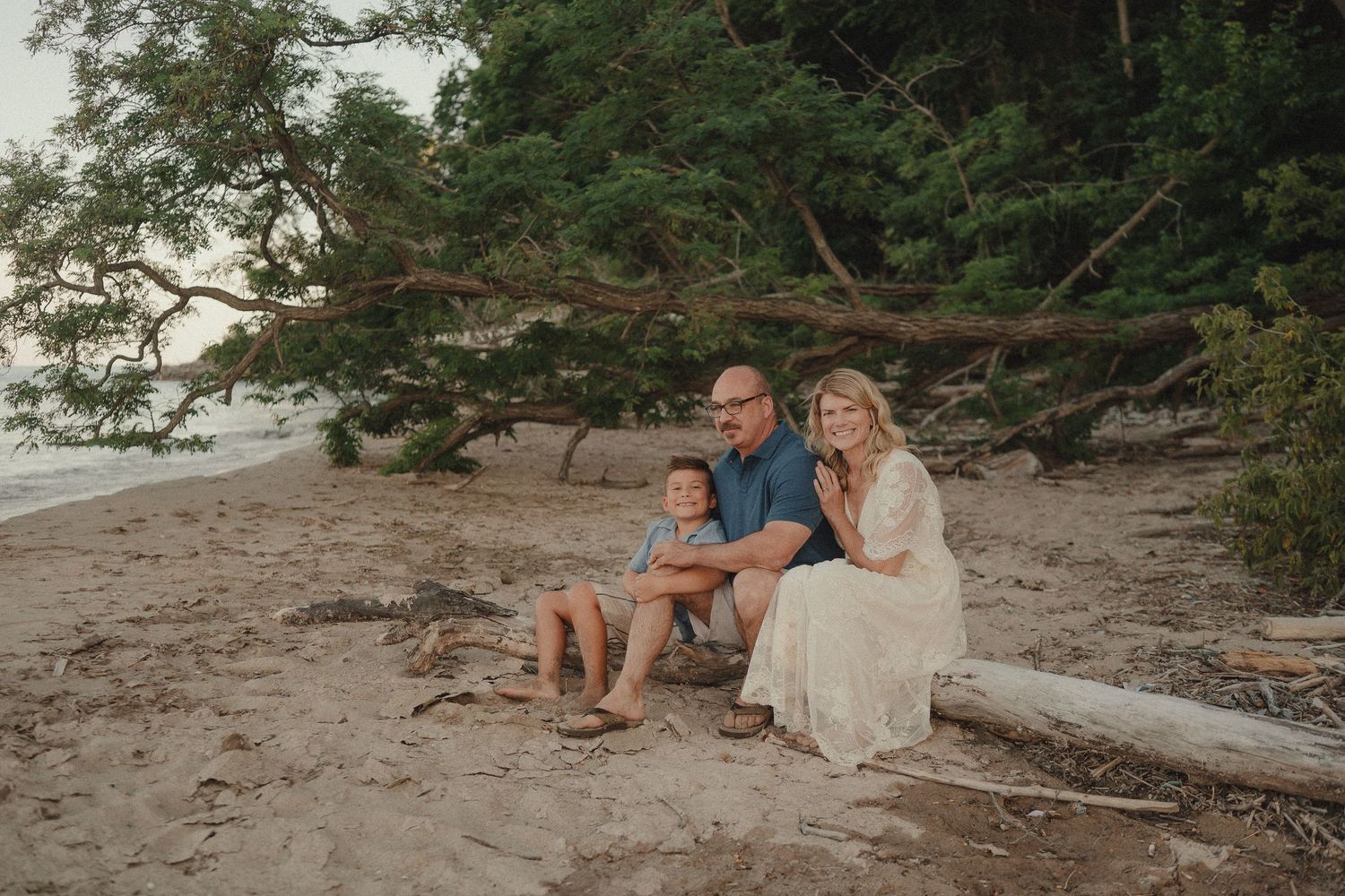 A happy family of three sits together on driftwood at a serene sandy beach surrounded by green trees.