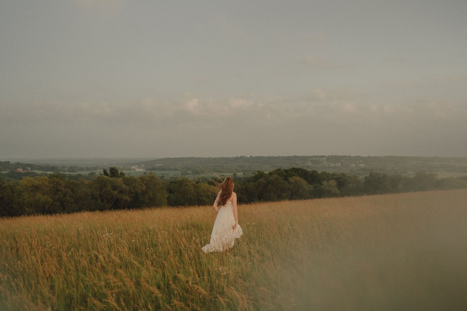 Young woman in a white dress stands in a golden wheat field under a dramatic overcast sky.