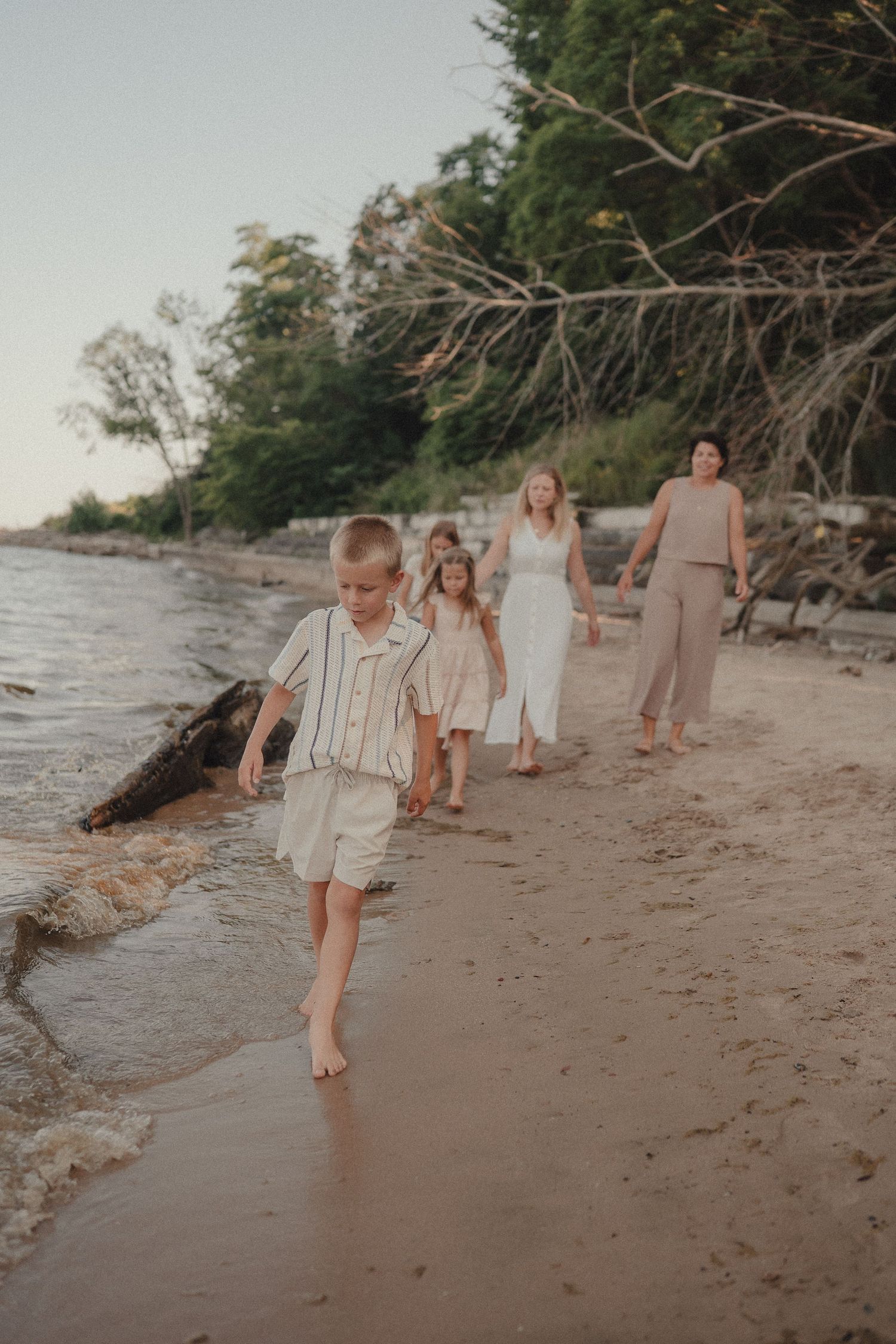 Family walking barefoot along sandy shoreline at dusk with trees in background.