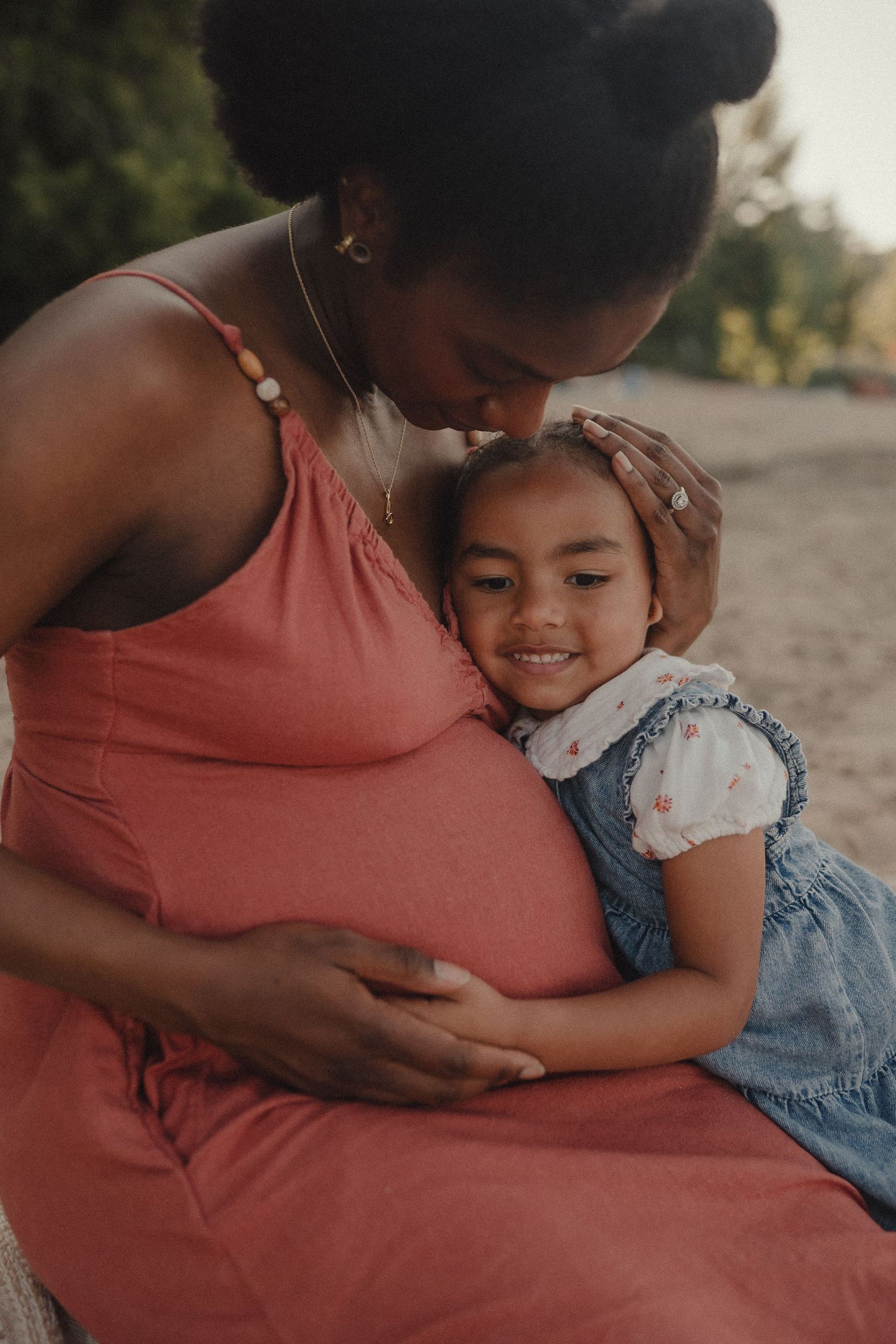 Pregnant woman in pink dress tenderly embraces smiling young girl outdoors in warm golden light.