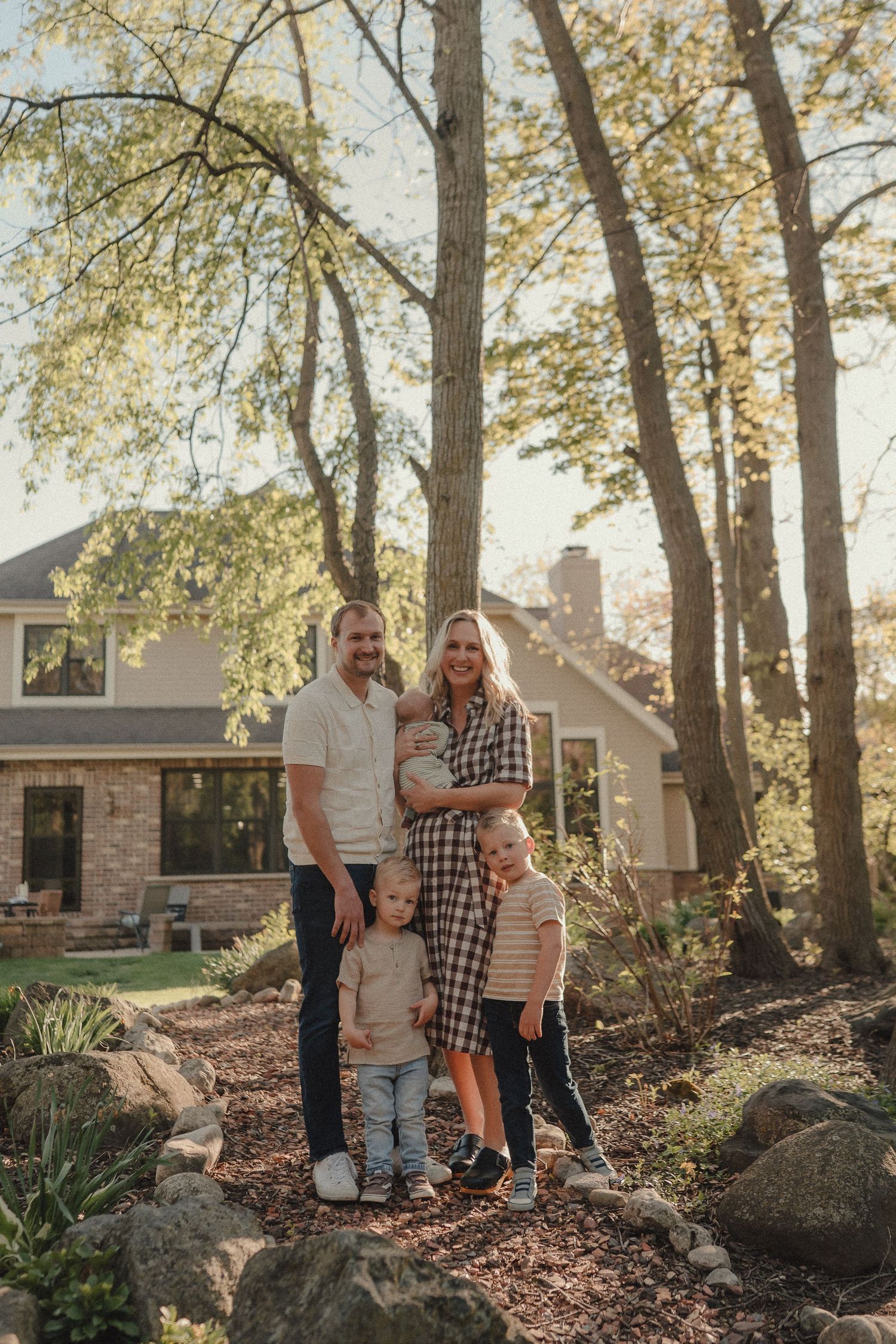 Happy family of five posing outdoors in golden hour light with brick house and tall trees behind them.