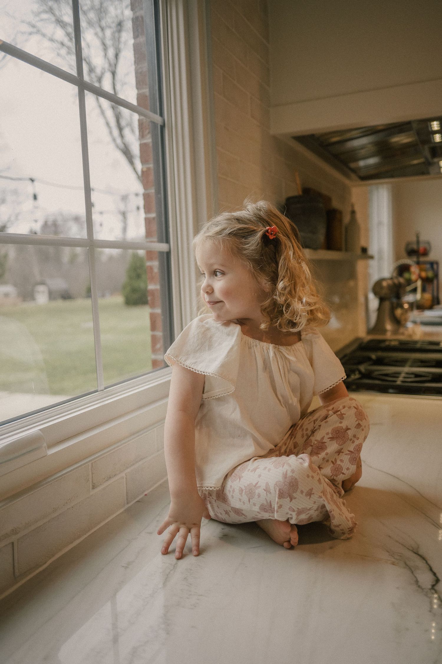 Curly-haired toddler girl in a white top sitting on a marble kitchen counter by a window.